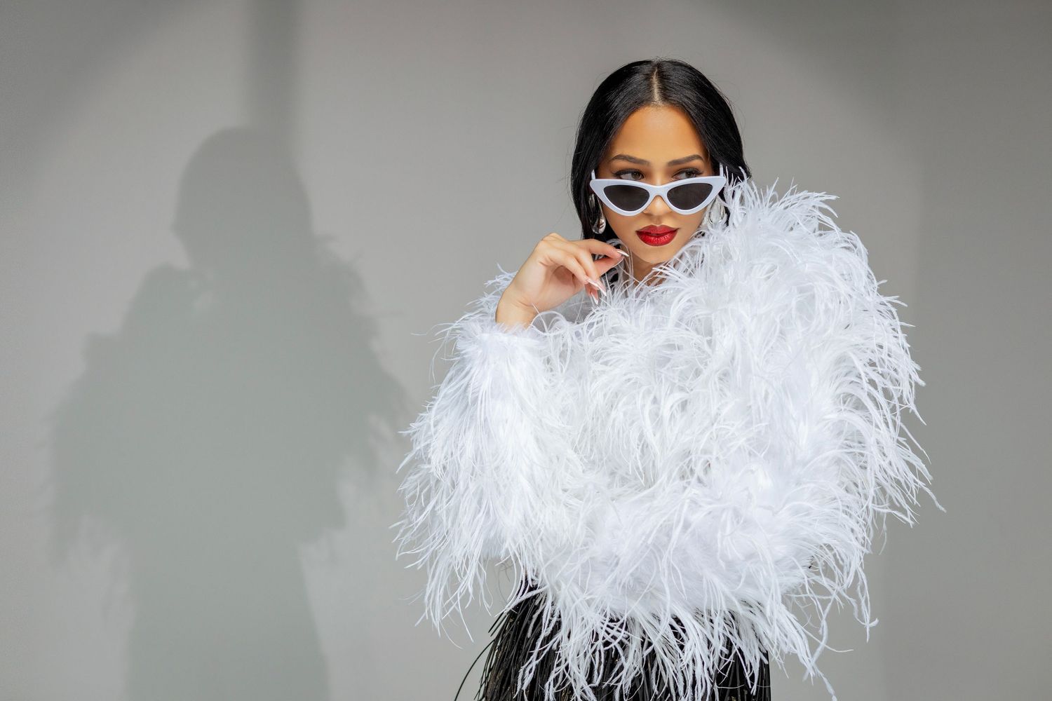 Fashionable model in retro sunglasses and fluffy white feathered jacket poses against gray wall.