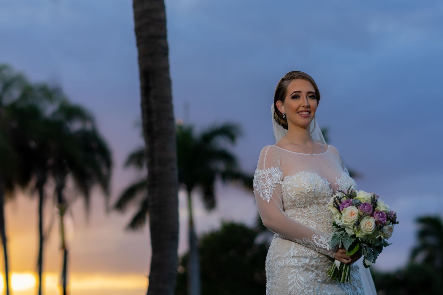 A bride holds a bouquet against a sunset sky with palm trees silhouetted in the background.