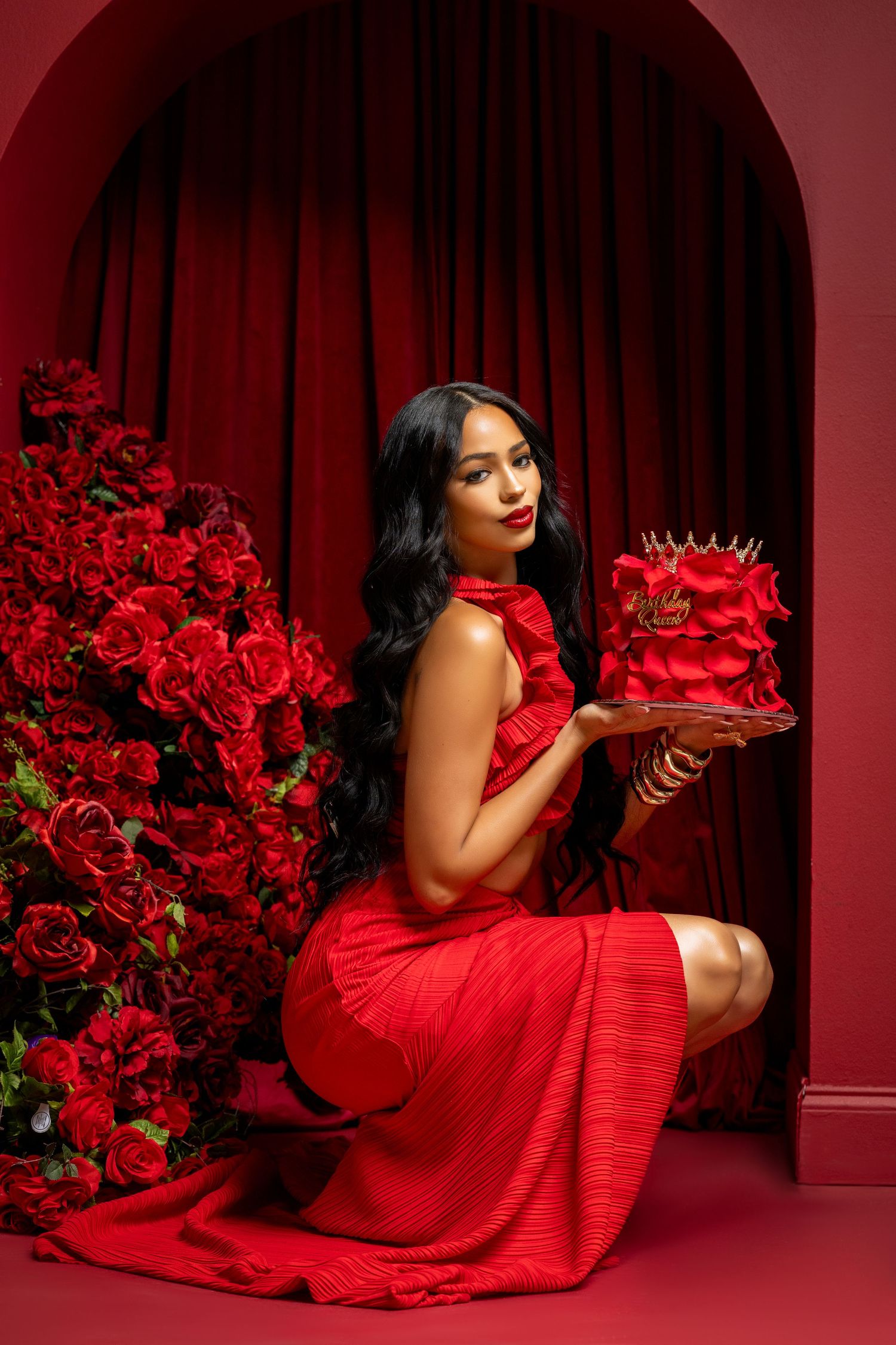 Woman in red dress holds elegant red crown cake while posing against red curtain backdrop with rose decorations.