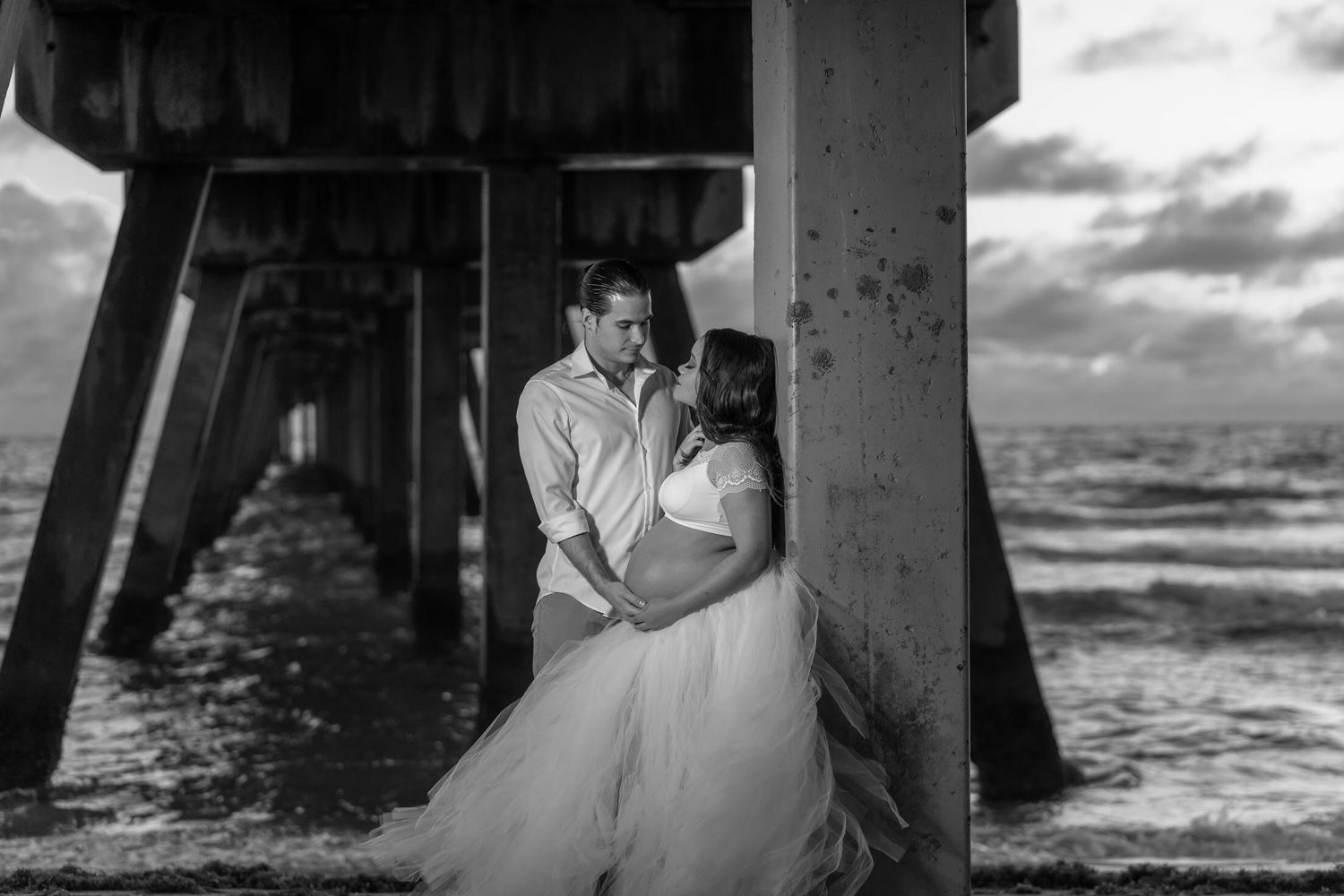 A wedding couple shares a romantic kiss under a pier by the ocean during sunset.