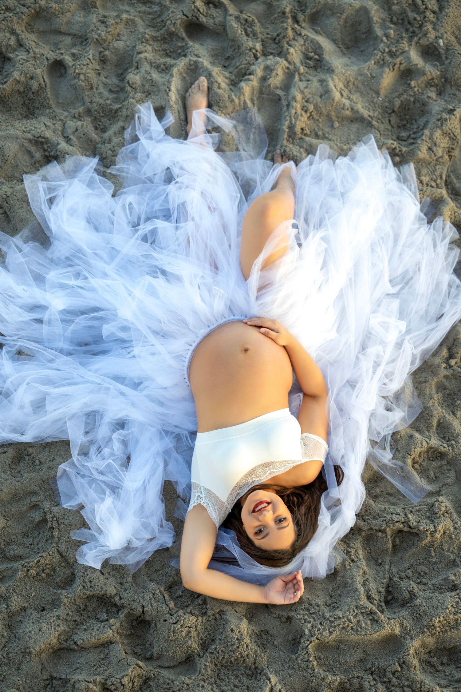 Expecting mother in white dress lies on sandy beach surrounded by flowing tulle fabric creating dreamy maternity photo.