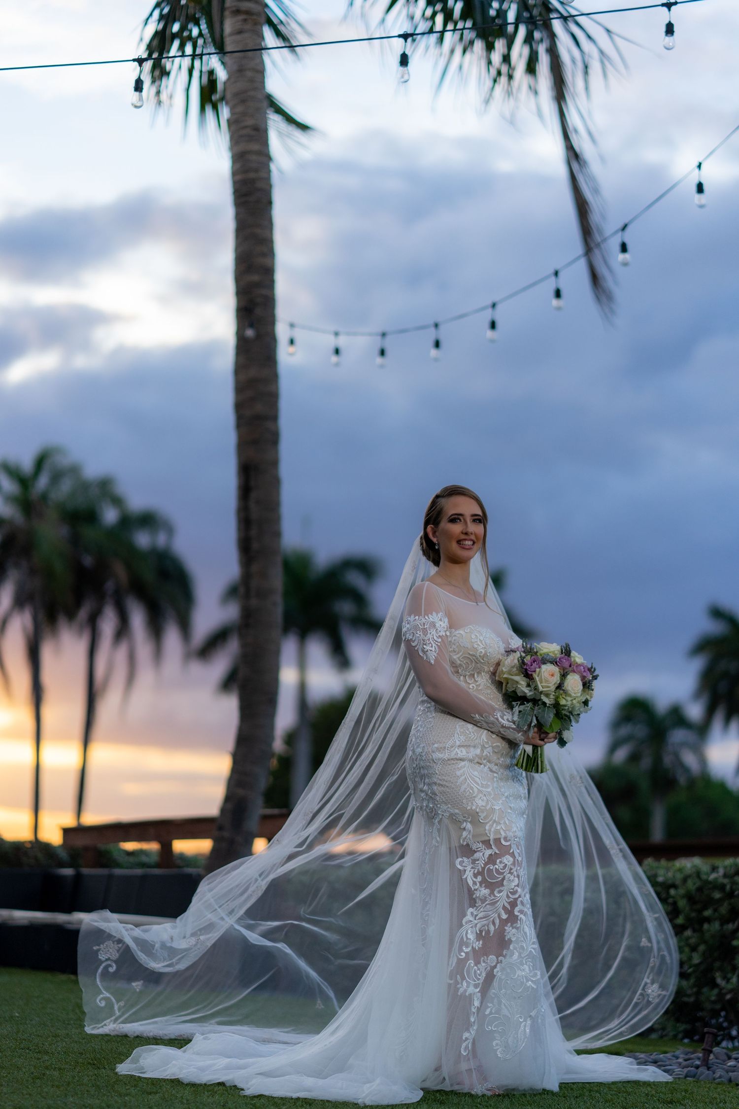 A bride in a flowing lace gown and veil poses on a lawn at sunset with palm trees and string lights in the background.