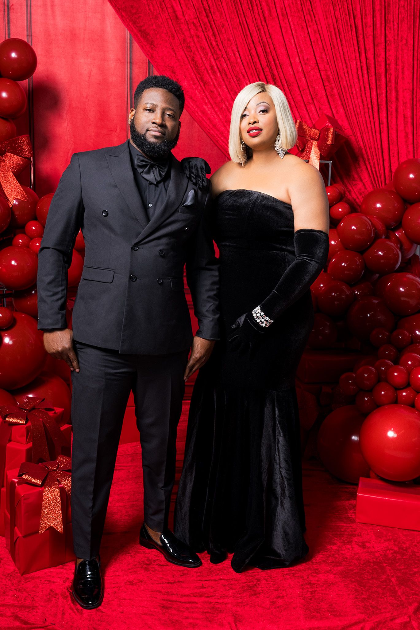 Formal couple posing against red backdrop with black formal attire and festive balloon decorations.