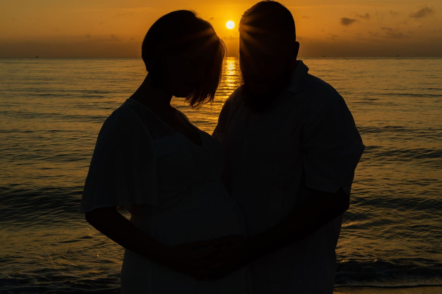 Silhouetted figures embrace on beach at sunset with golden light reflecting off ocean waves in romantic scene.