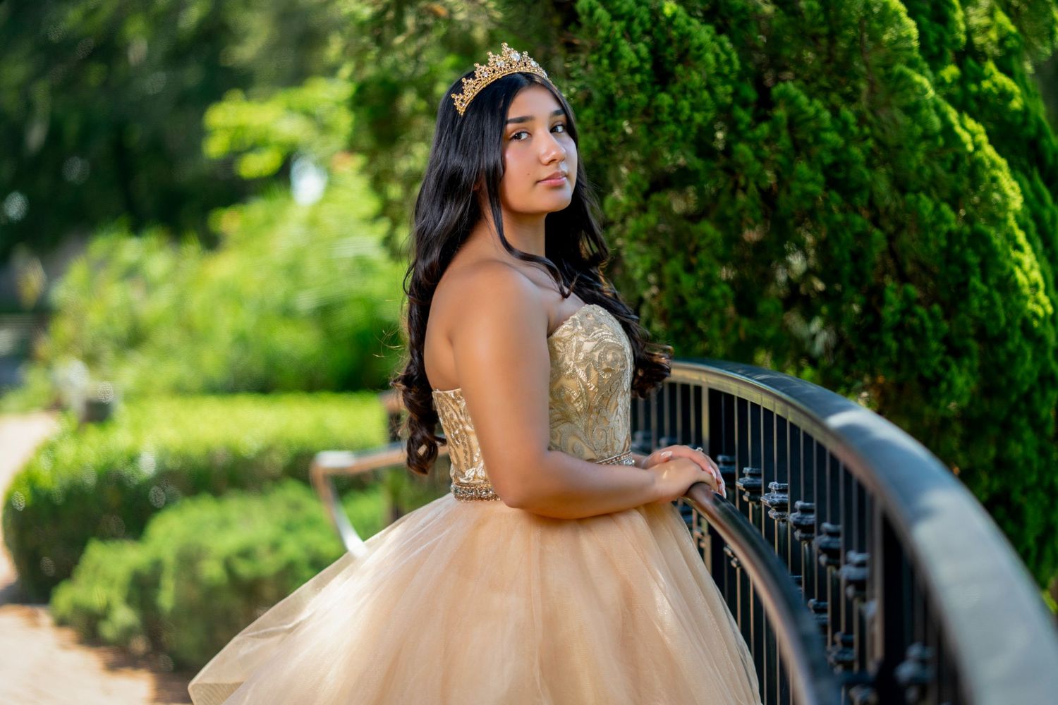 A young woman in a formal gown poses in a lush garden setting with an ornate wrought iron railing.