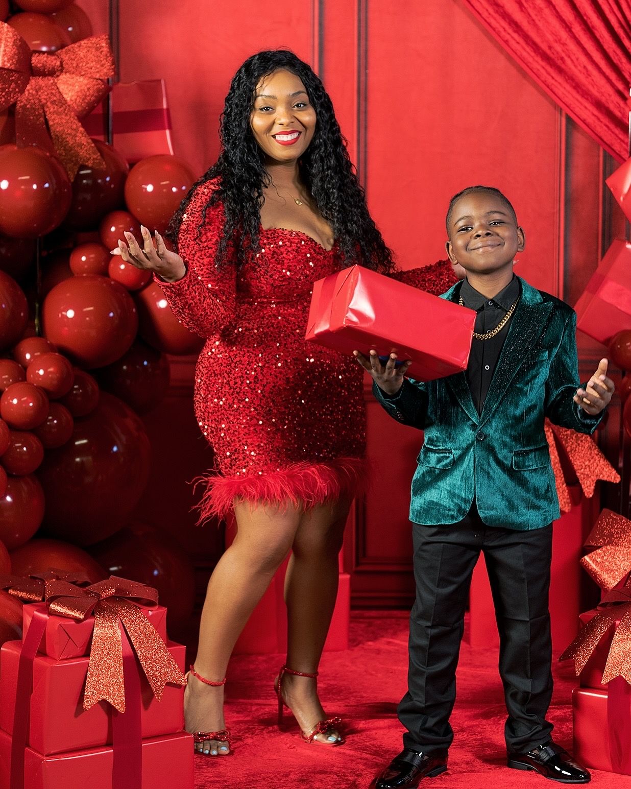 Festive holiday photo featuring red sequin dress and green velvet blazer against backdrop of red balloons and wrapped gifts.
