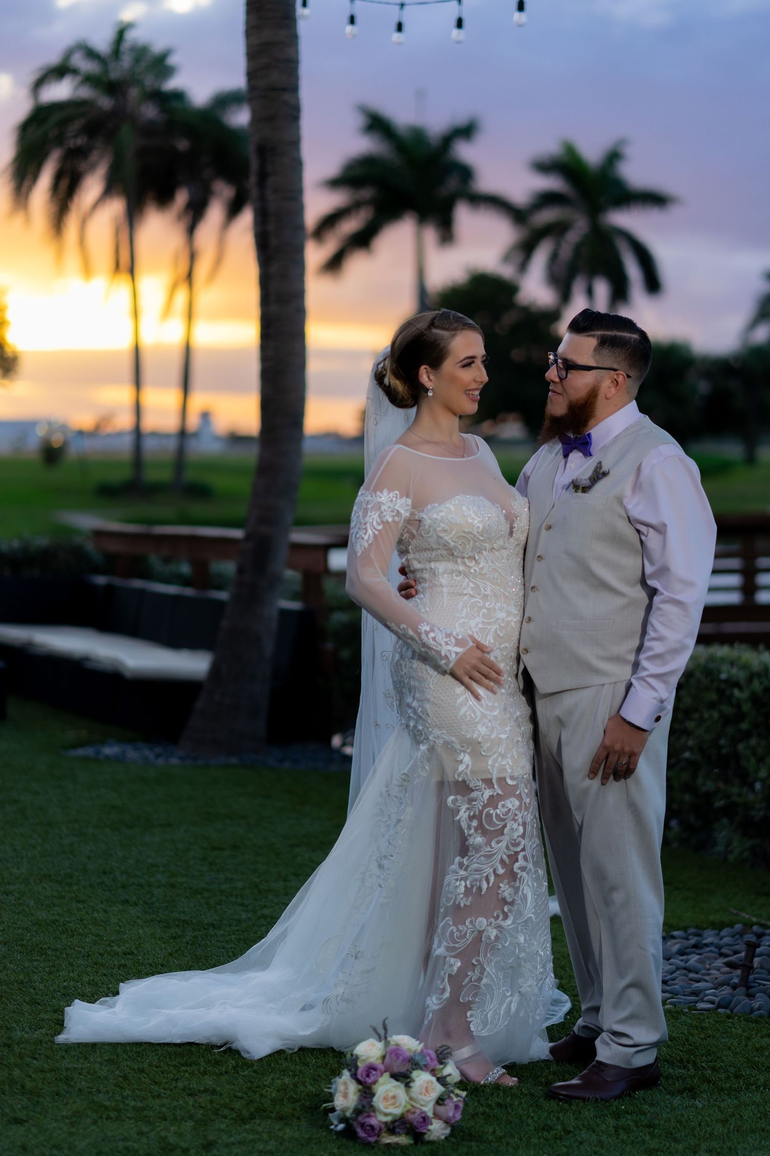 A romantic sunset wedding portrait with palm trees silhouetted against the colorful sky on a tropical beach lawn.
