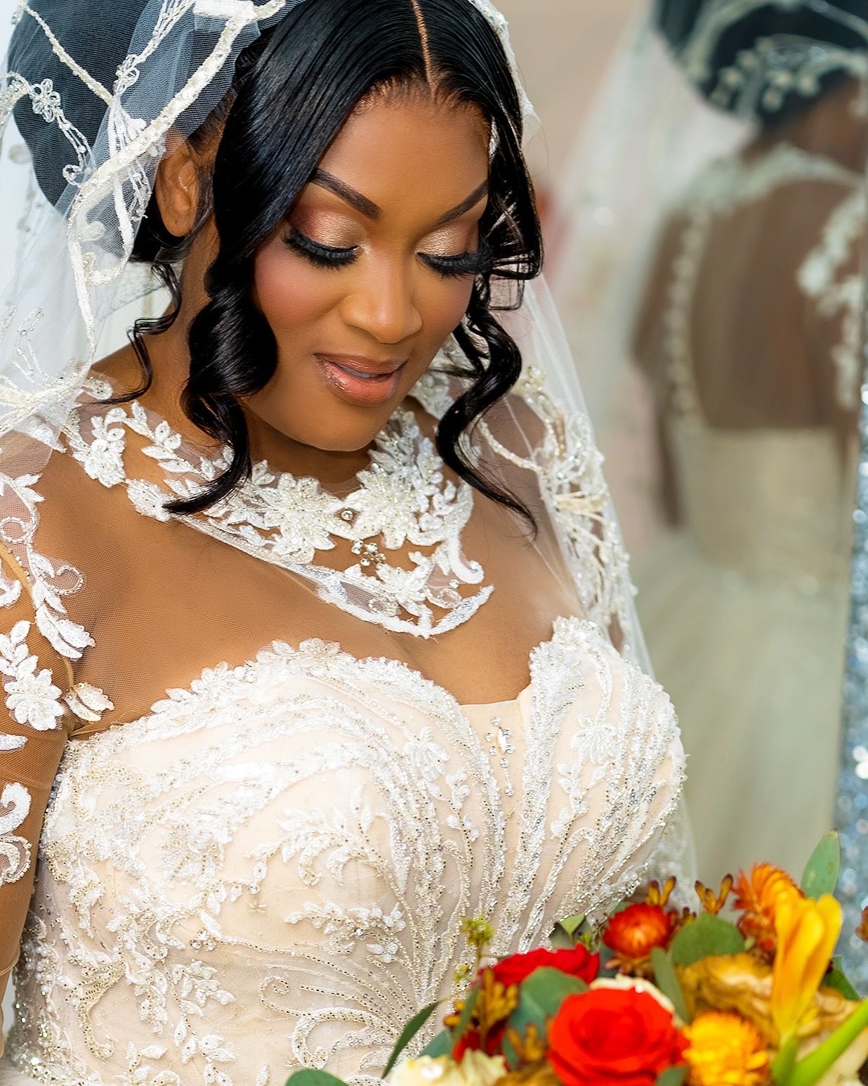 Bride wearing an elegant white lace wedding dress with sheer details holds a colorful bouquet with red and yellow flowers.