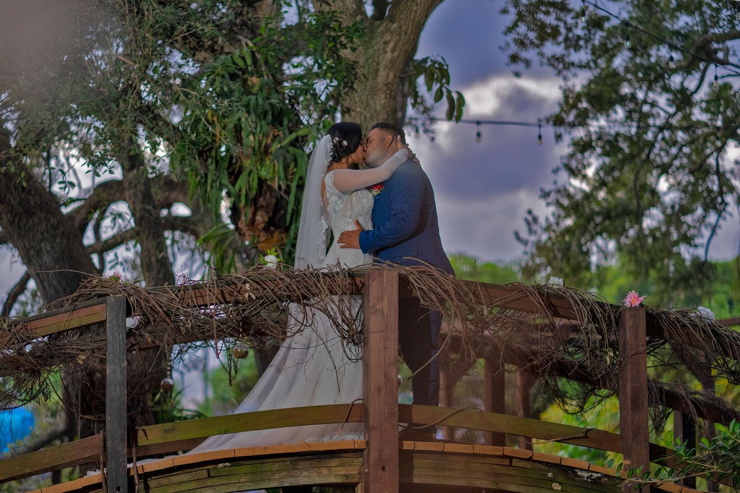 A couple embraces on a rustic wooden bridge surrounded by lush green trees at sunset.