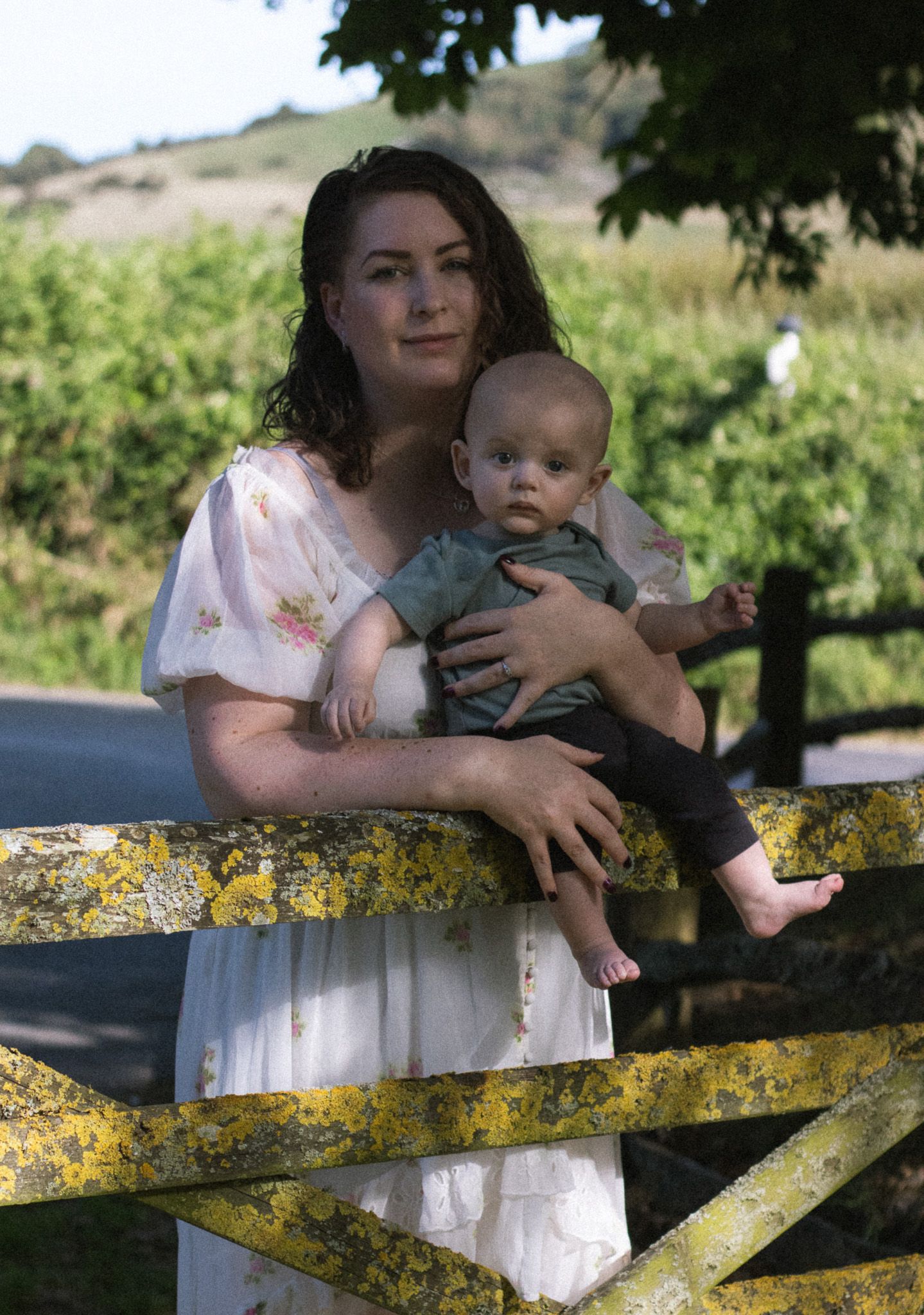 A baby in a white dress sits on a yellow lichen-covered wooden fence on a sunny day.