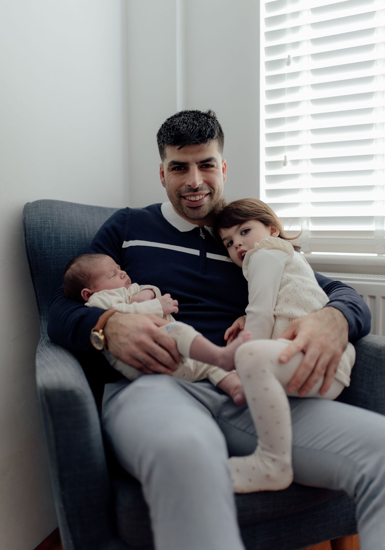 A sequence of tender moments showing a parent cradling and bonding with a newborn babying a toddler on a grey couch by a window.