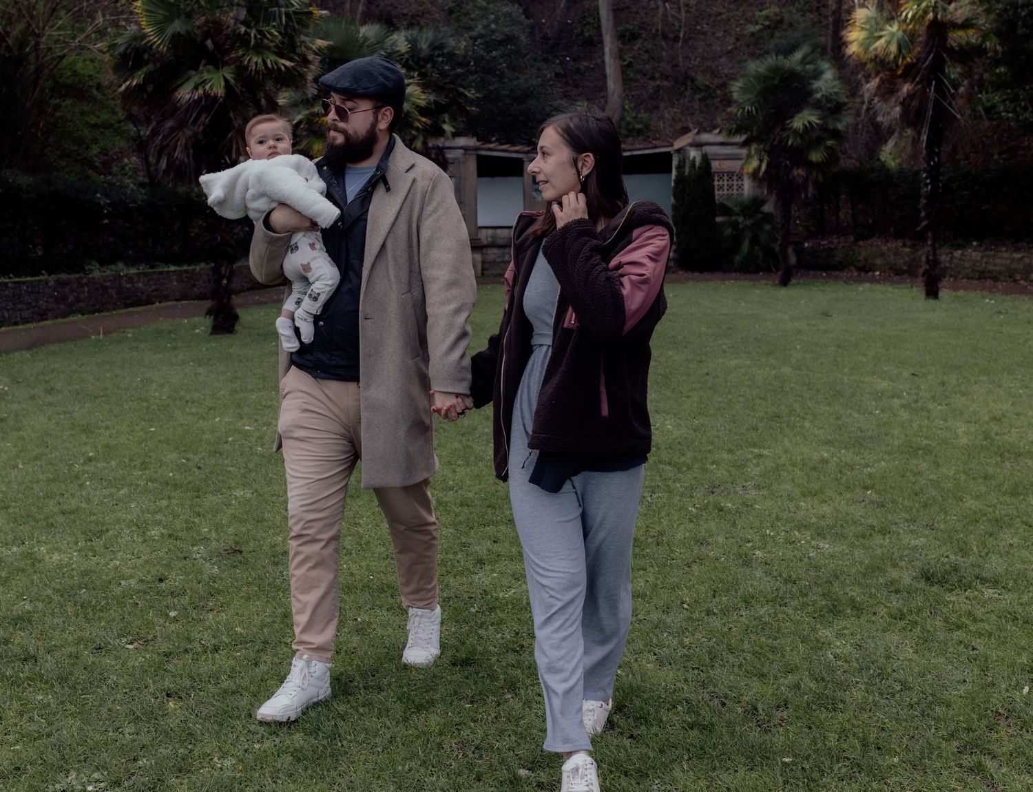 A couple walks together on a grassy lawn while each holding a small baby in their arms.