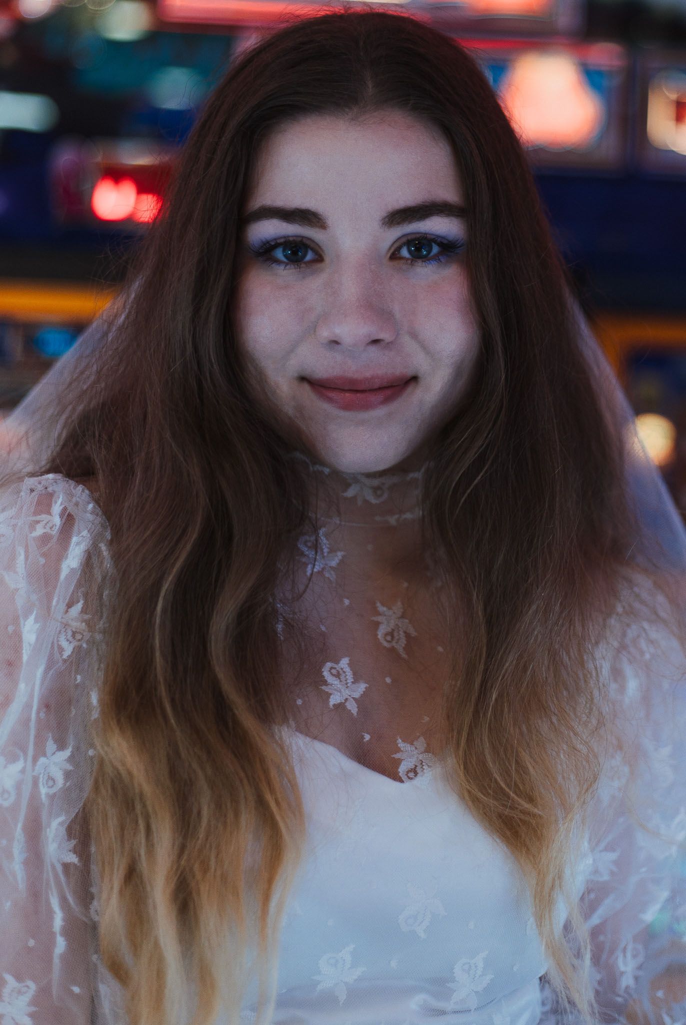 Bride at an arcade poses playfully in a white dress with ombre hair against neon signs.