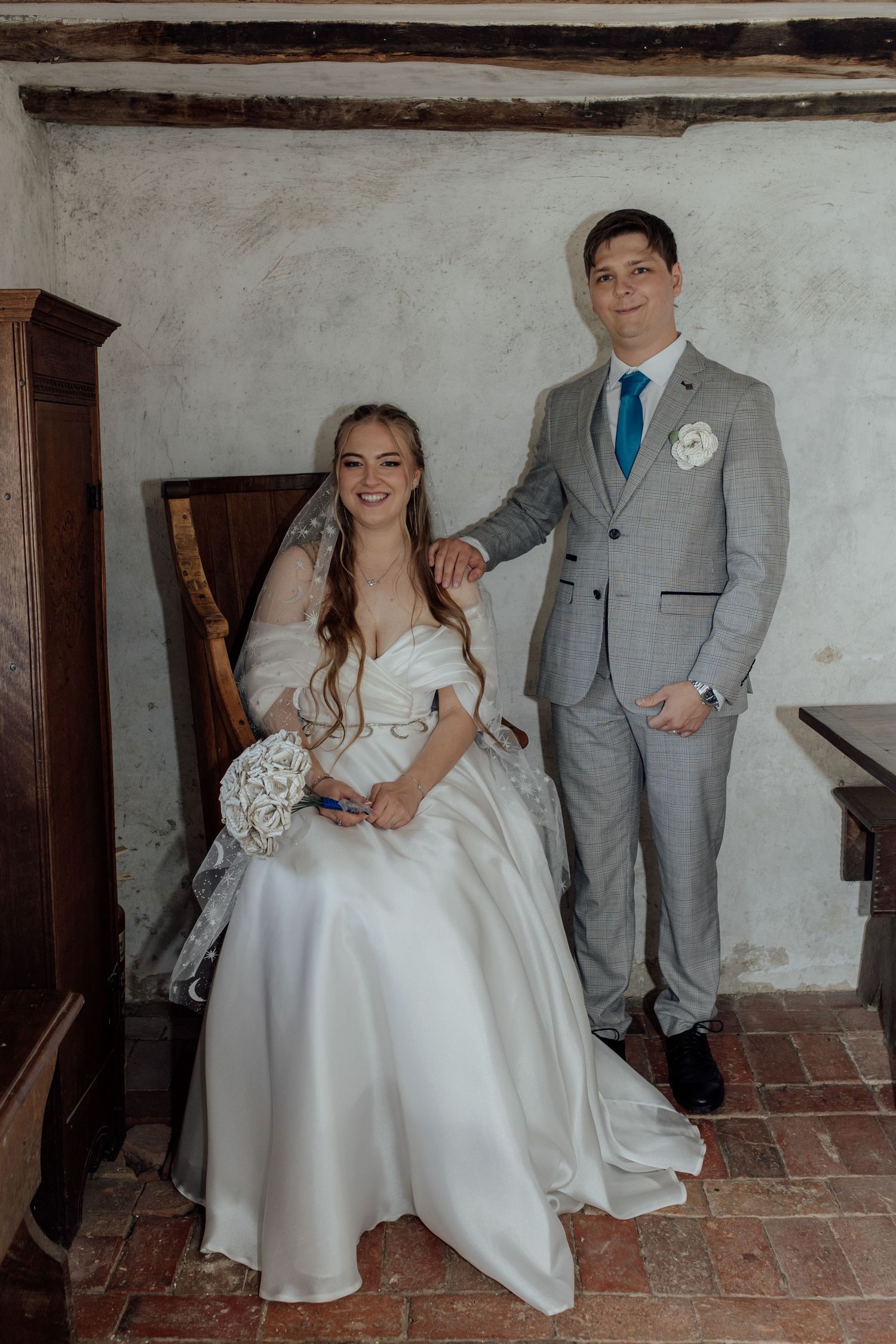 Newlyweds posing against white wall with exposed wooden beams in elegant attire.