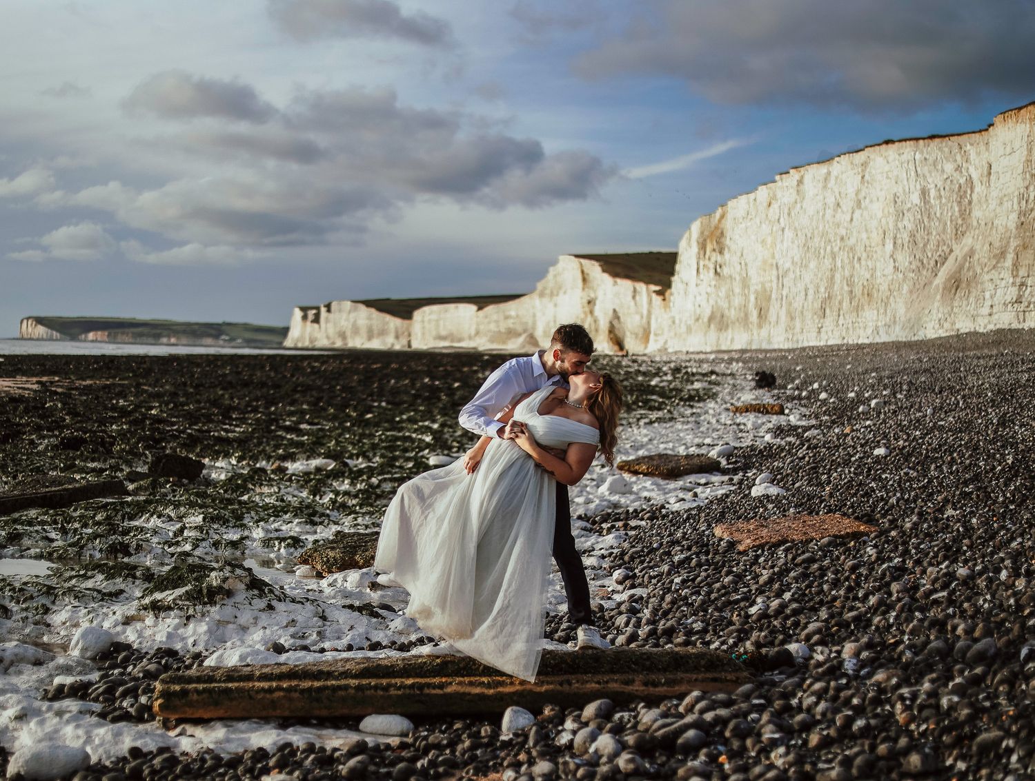 A couple embraces on a rocky beach with dramatic white chalk cliffs of Seven Sisters, Sussex Coast in the background.