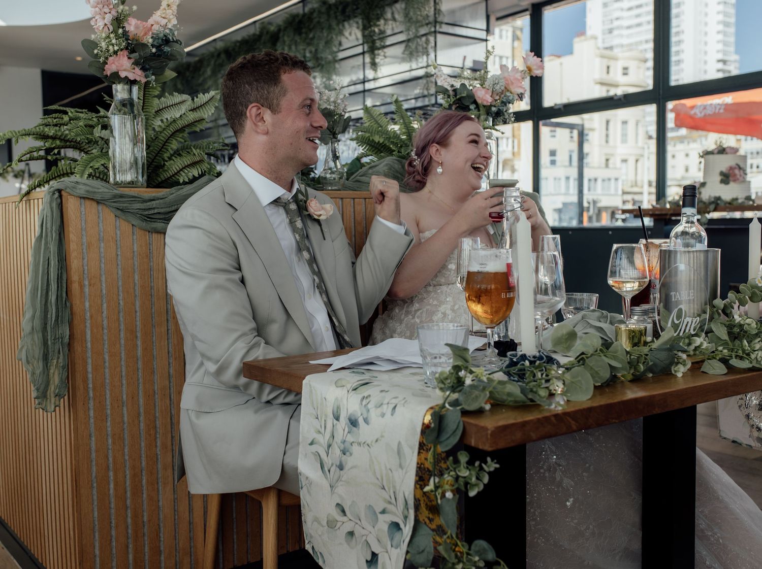 A couple in wedding attire share a drink at a restaurant bar decorated with greenery and flowers.