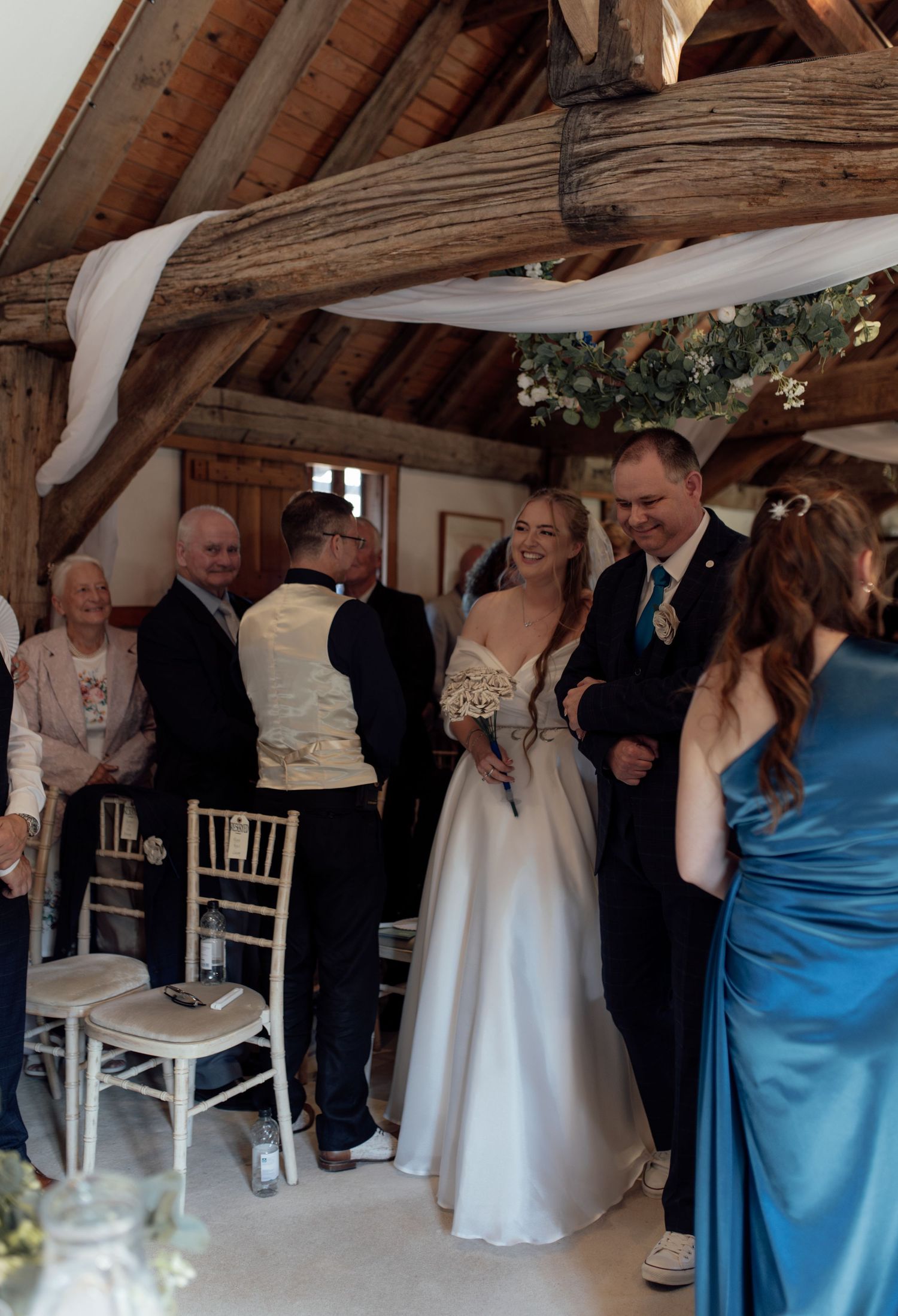 Wedding guests gather in a rustic wooden barn venue with exposed beams and white chairs for an indoor ceremony.
