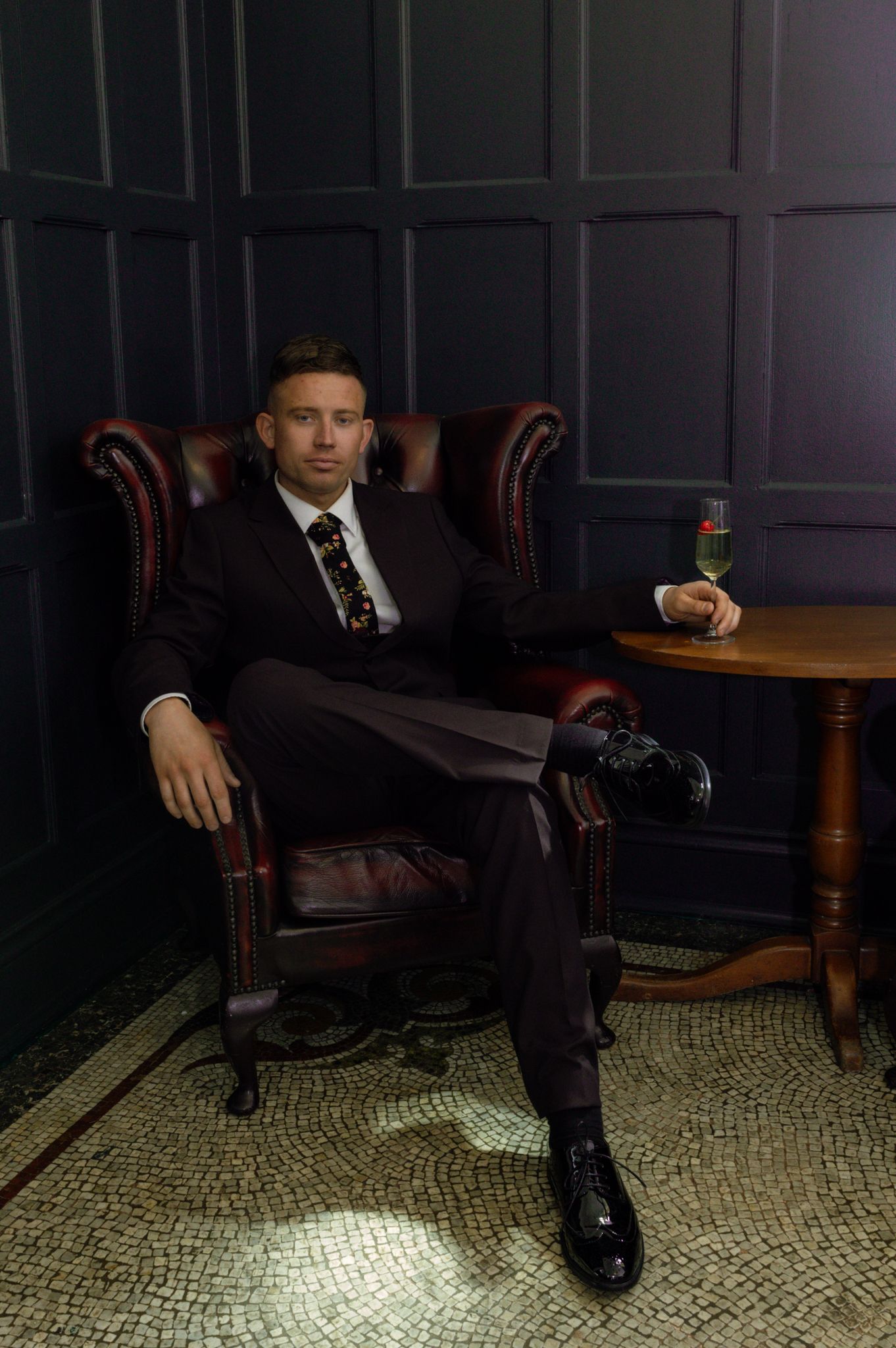 Person in formal attire sits in dark moody lighting on vintage leather chair.