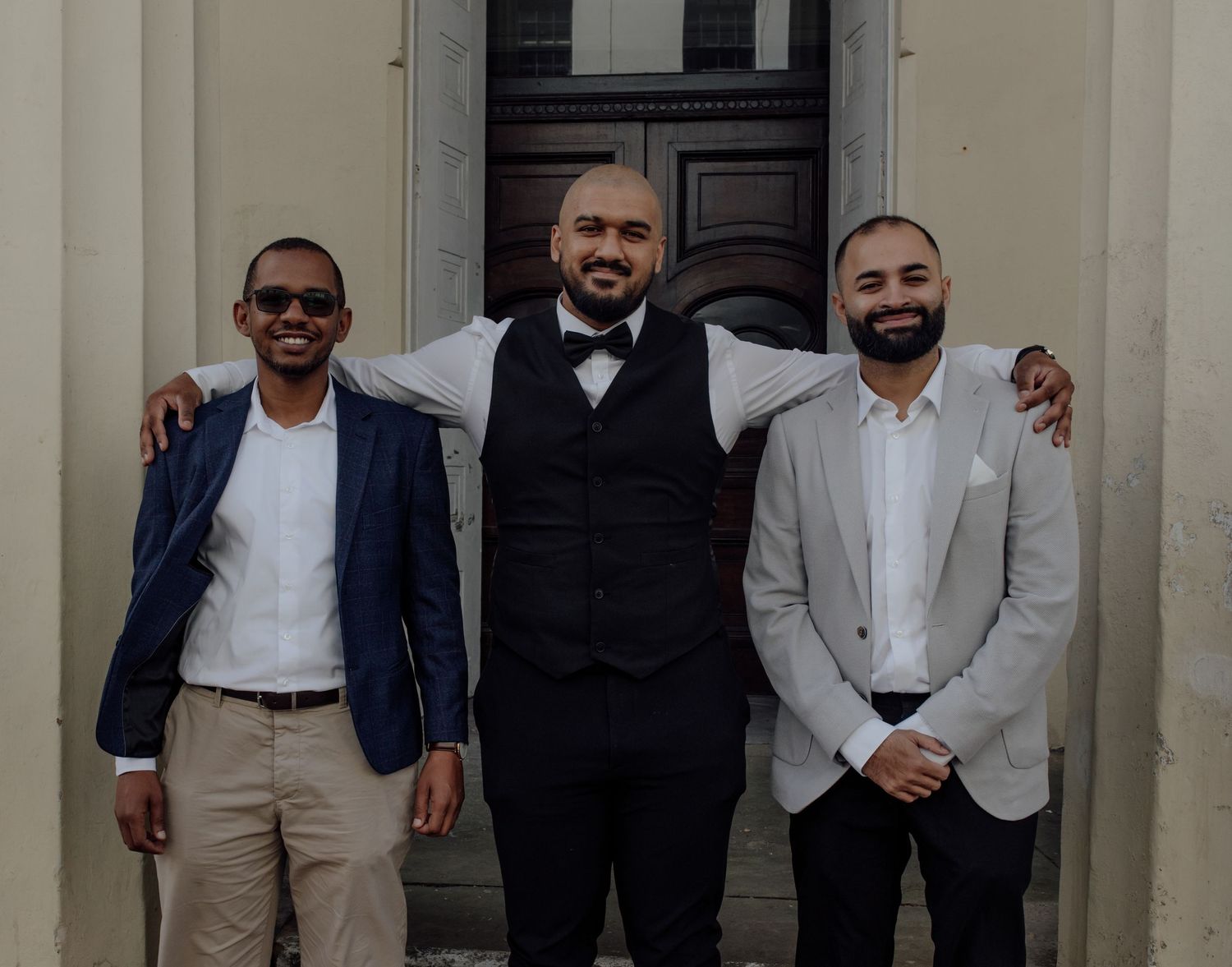 A group of well-dressed men in suits and formal attire pose together against a light-colored wall.