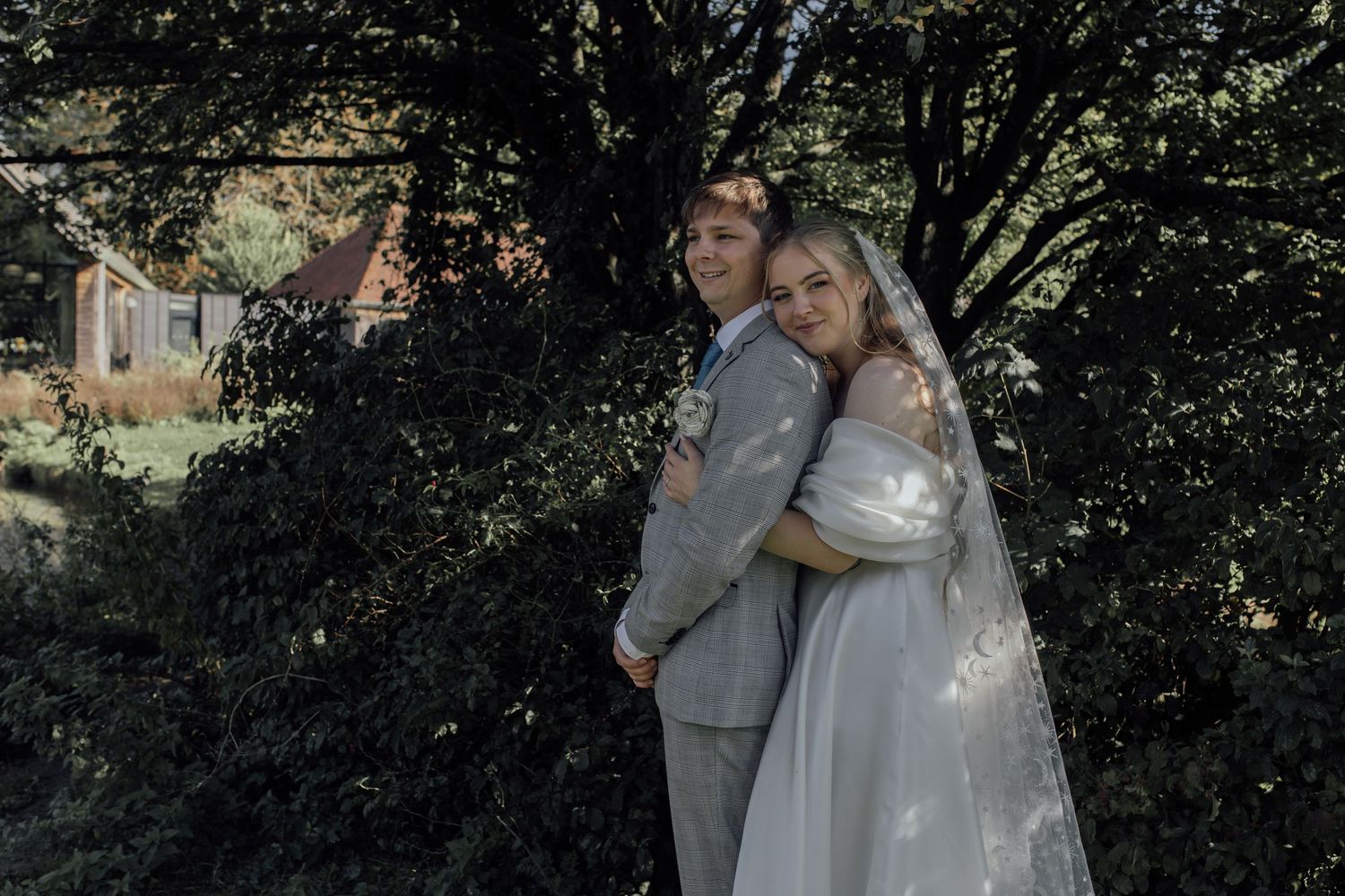 A couple shares a romantic moment under lush green trees during their outdoor wedding celebration.