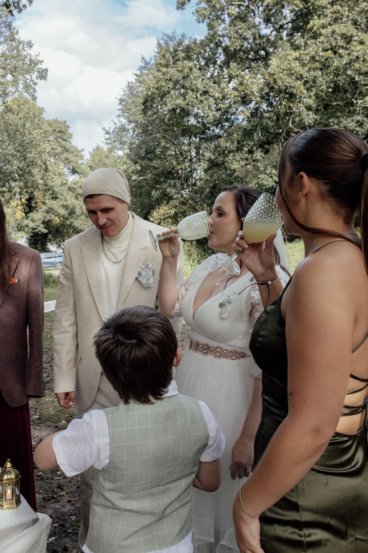 A group gathers outdoors under trees for a casual wedding ceremony in natural lighting.