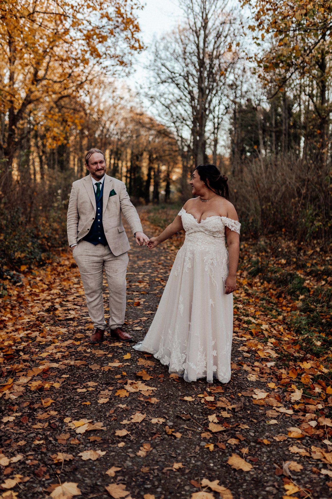 A couple holds hands on a leaf-covered path surrounded by autumn trees at their fall woodland wedding.