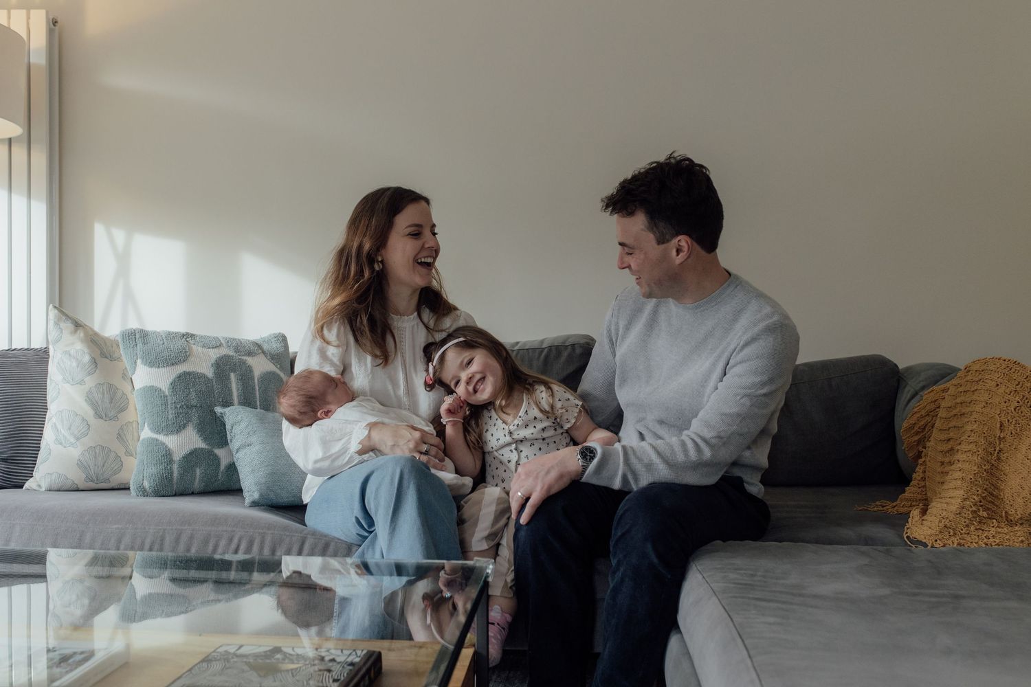 A family relaxes together on a gray couch in their bright living room with a yellow throw blanket.