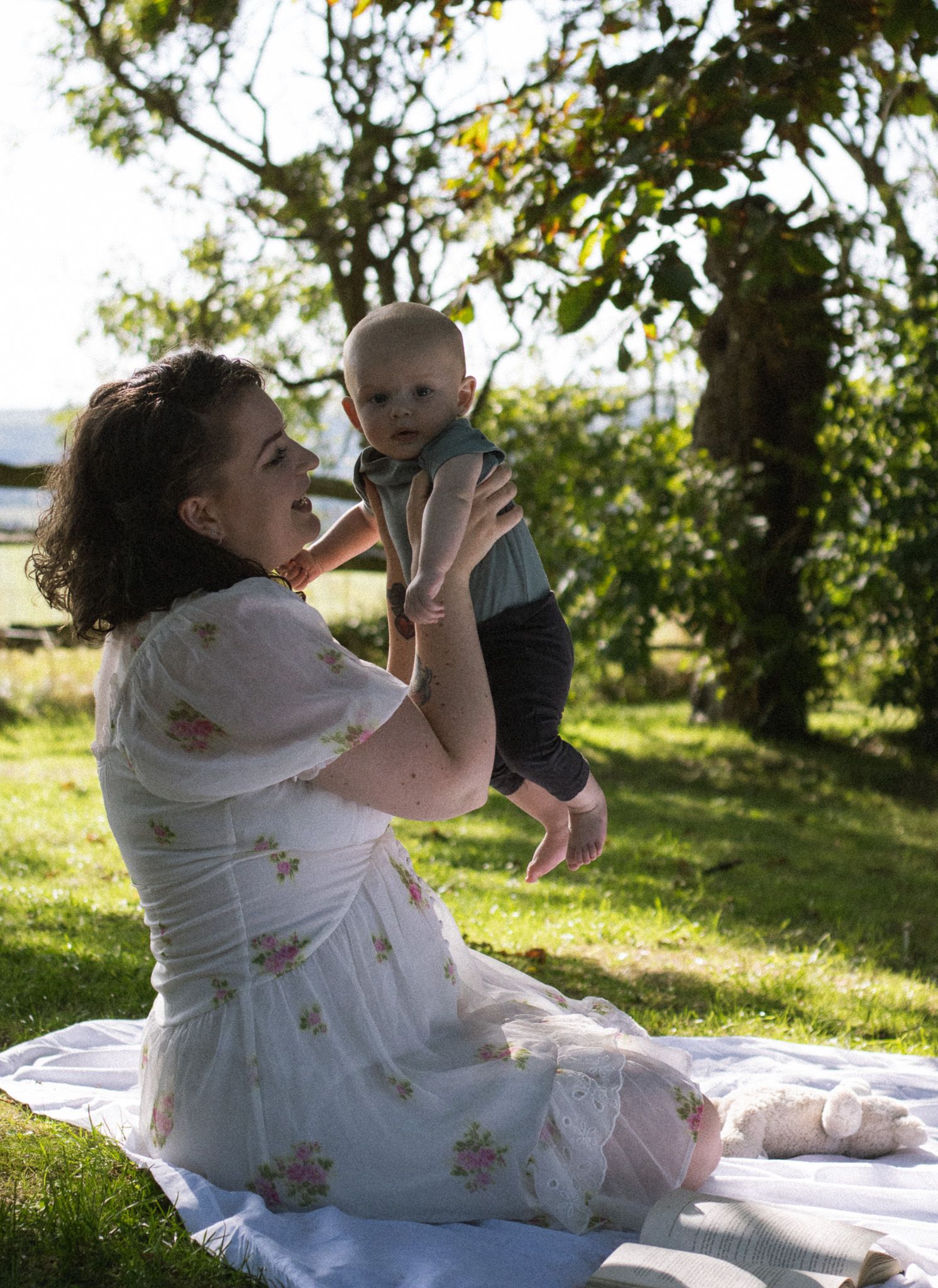 A person in a floral dress sits on a white blanket in a sunny orchard, living her son up.