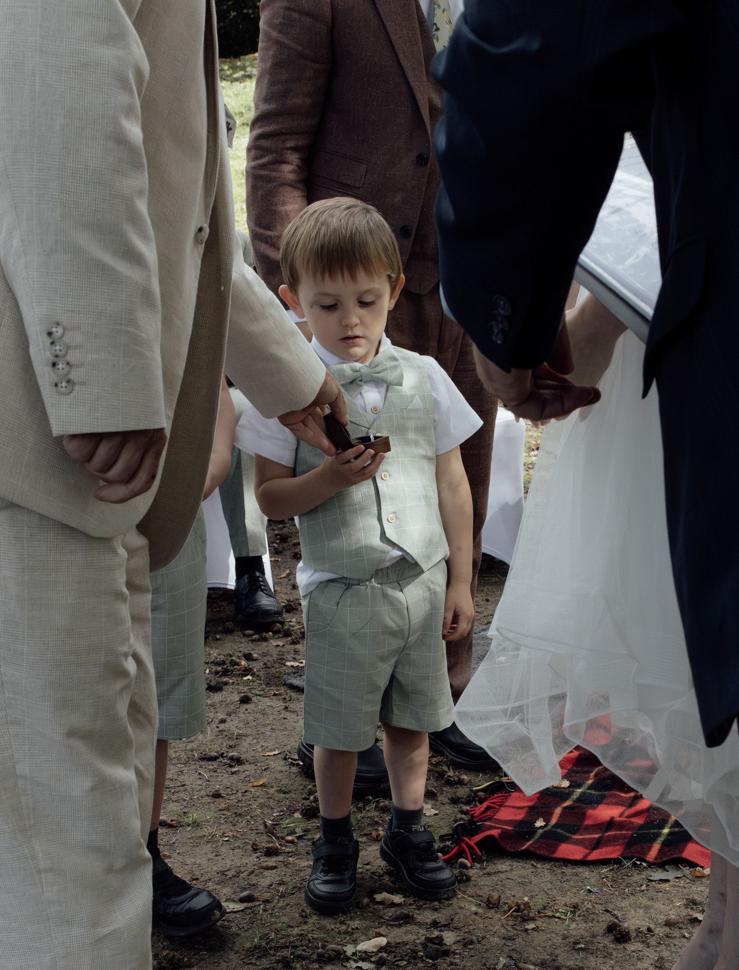 A child in khaki shorts stands between adults in white clothing on a dirt ground at an outdoor gathering.