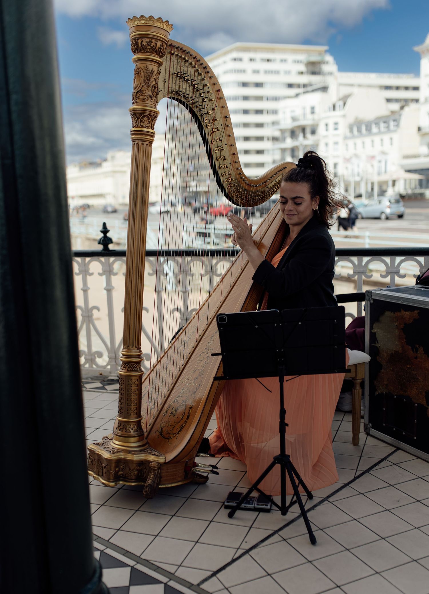A harpist performs on an outdoor terrace with white buildings and blue sky visible in the background.