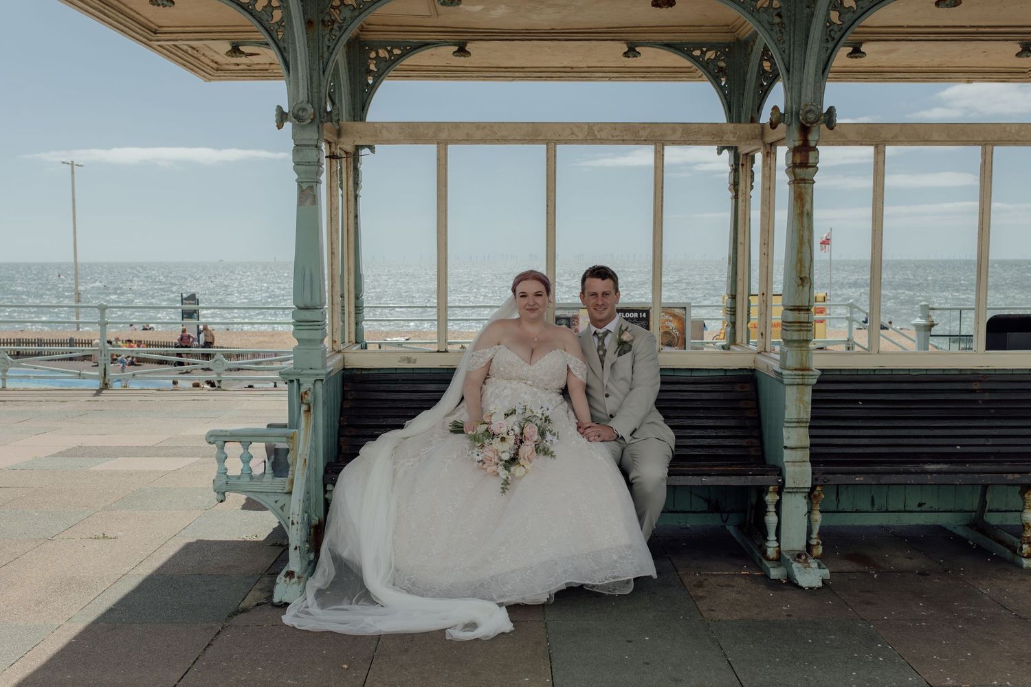 A bride and groom sit together on a bench at a beachfront gazebo during their coastal wedding ceremony.