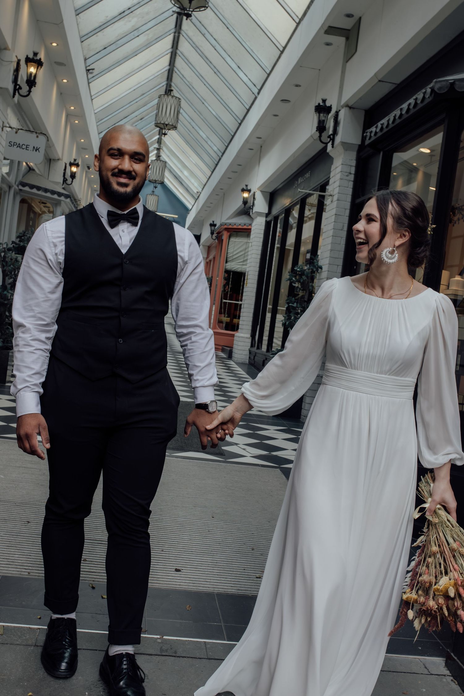 A couple in wedding attire walk hand in hand down a covered shopping arcade with white walls and black storefronts.