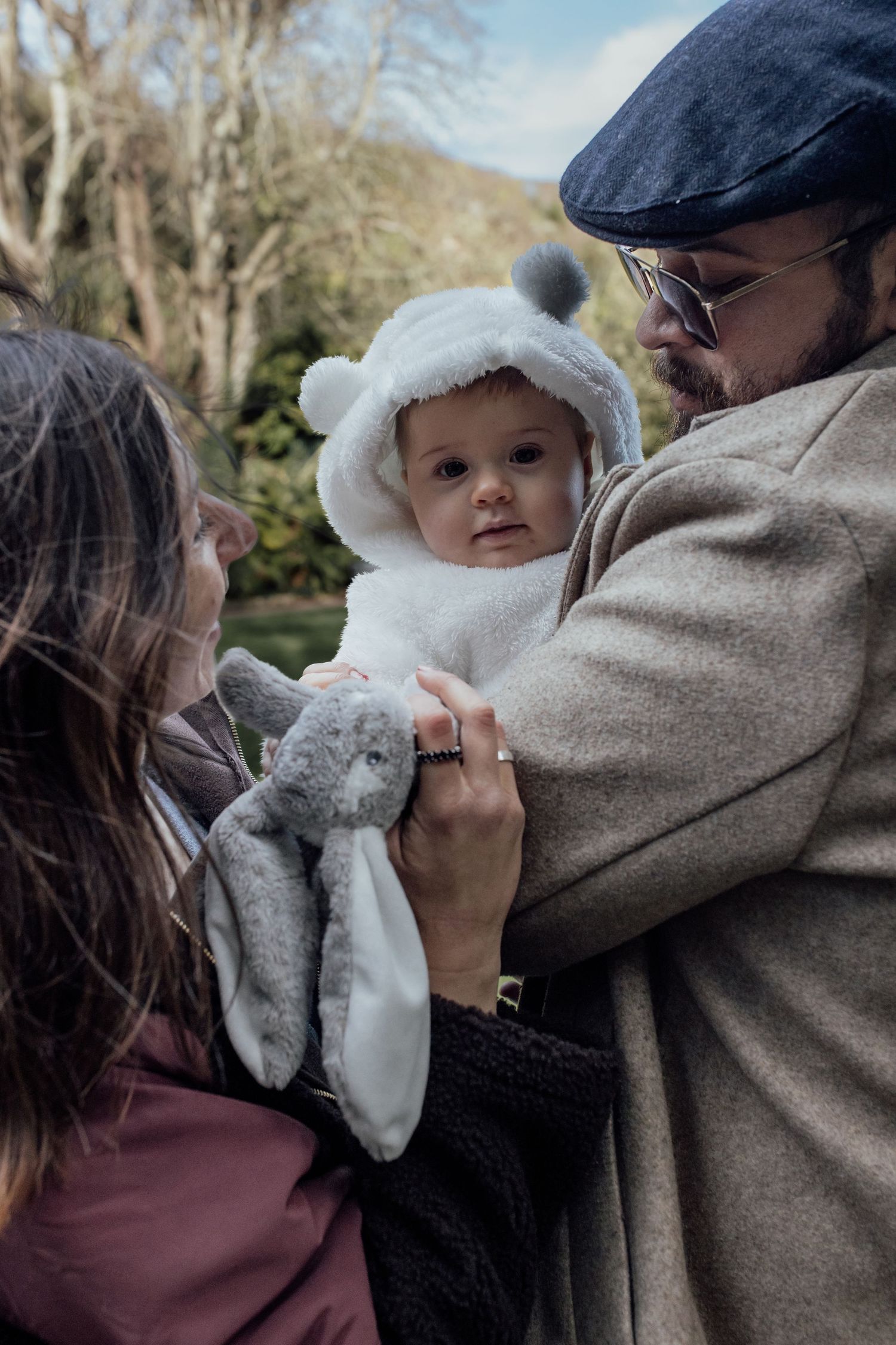 A baby wearing a white fuzzy winter hat is held outdoors while being shown a plush toy bunny on an autumn day.