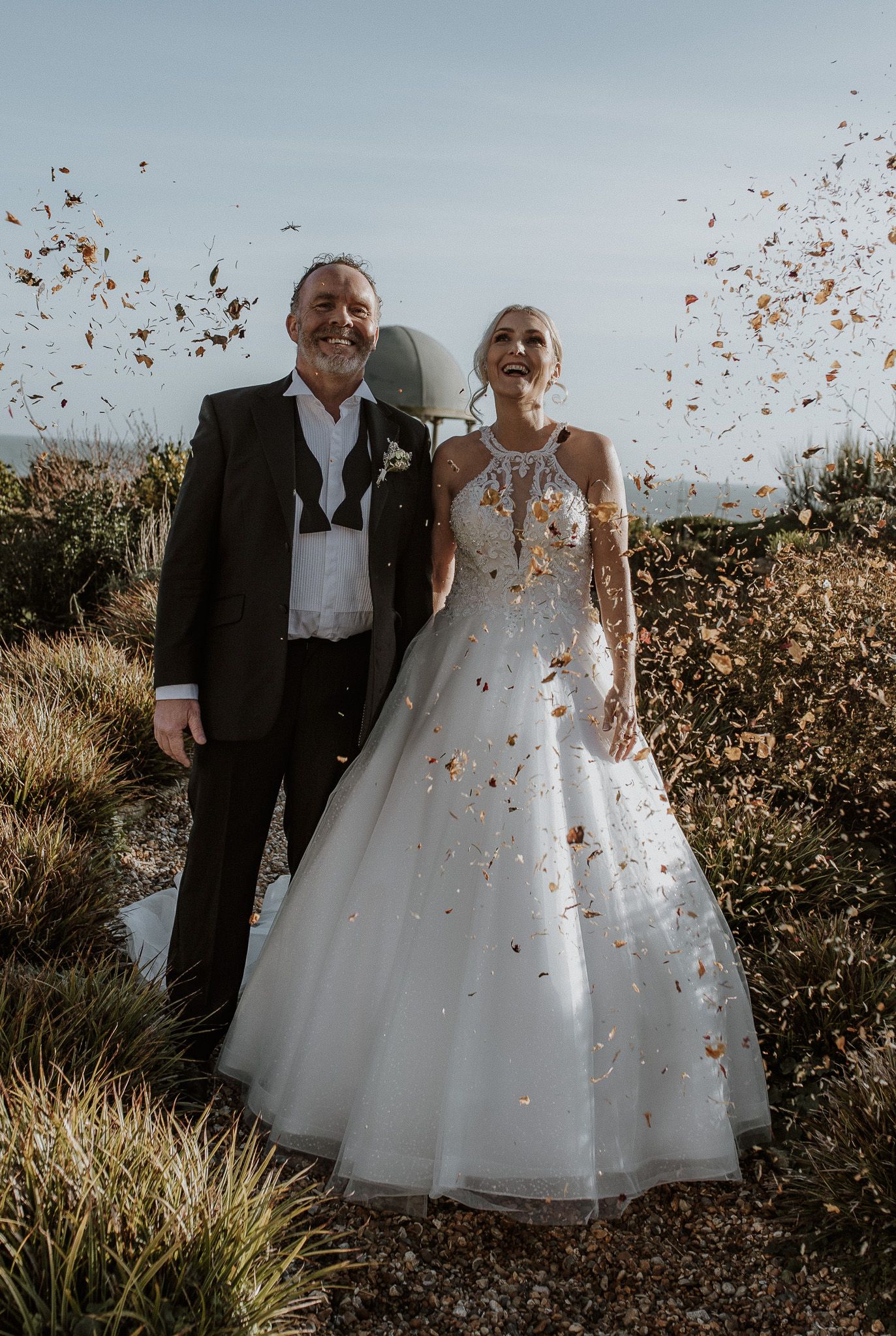 A romantic fall wedding photo with falling autumn leaves swirling around a couple in formal attire outdoors.