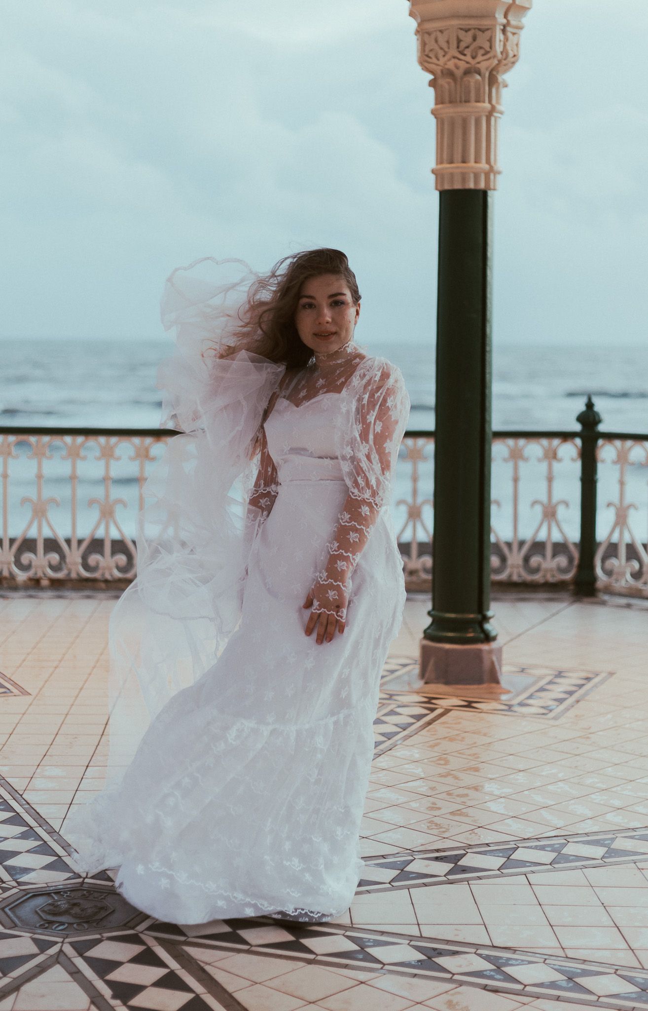 A bride in a flowing white lace dress stands on a tiled balcony overlooking the ocean as her hair blows in the wind.