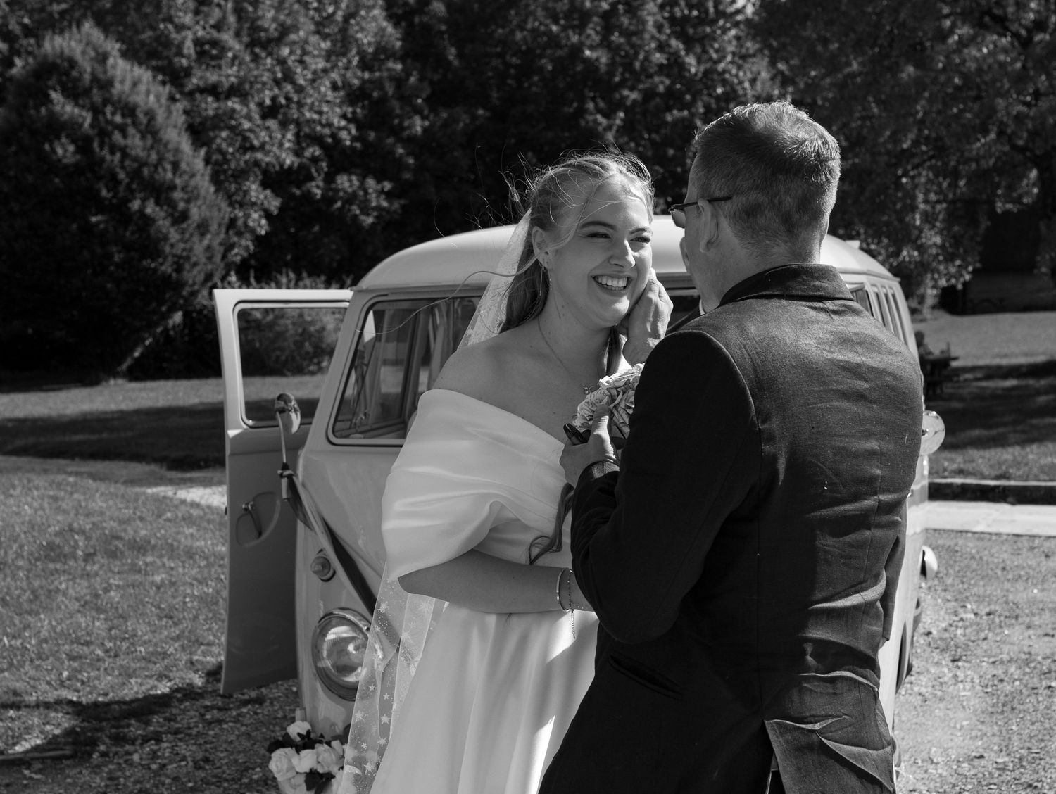 A black and white wedding photo with a classic car in the background on a sunny day.