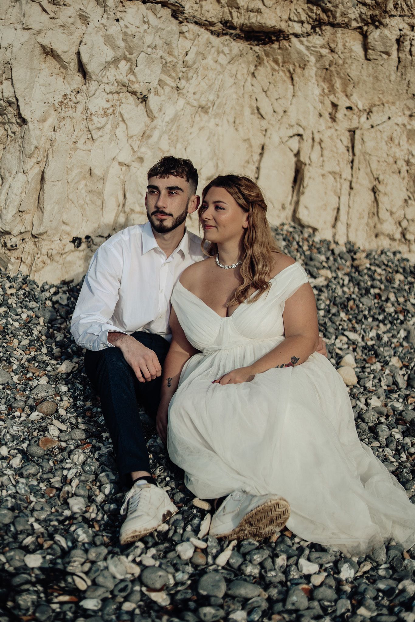 A couple sits together on rocky beach against a limestone cliff in romantic wedding attire.
