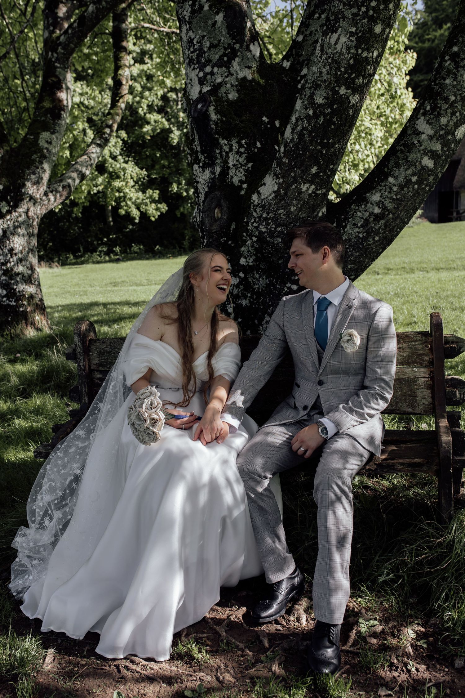 A couple in wedding attire shares a joyful moment while sitting together on a rustic bench under a shady tree.