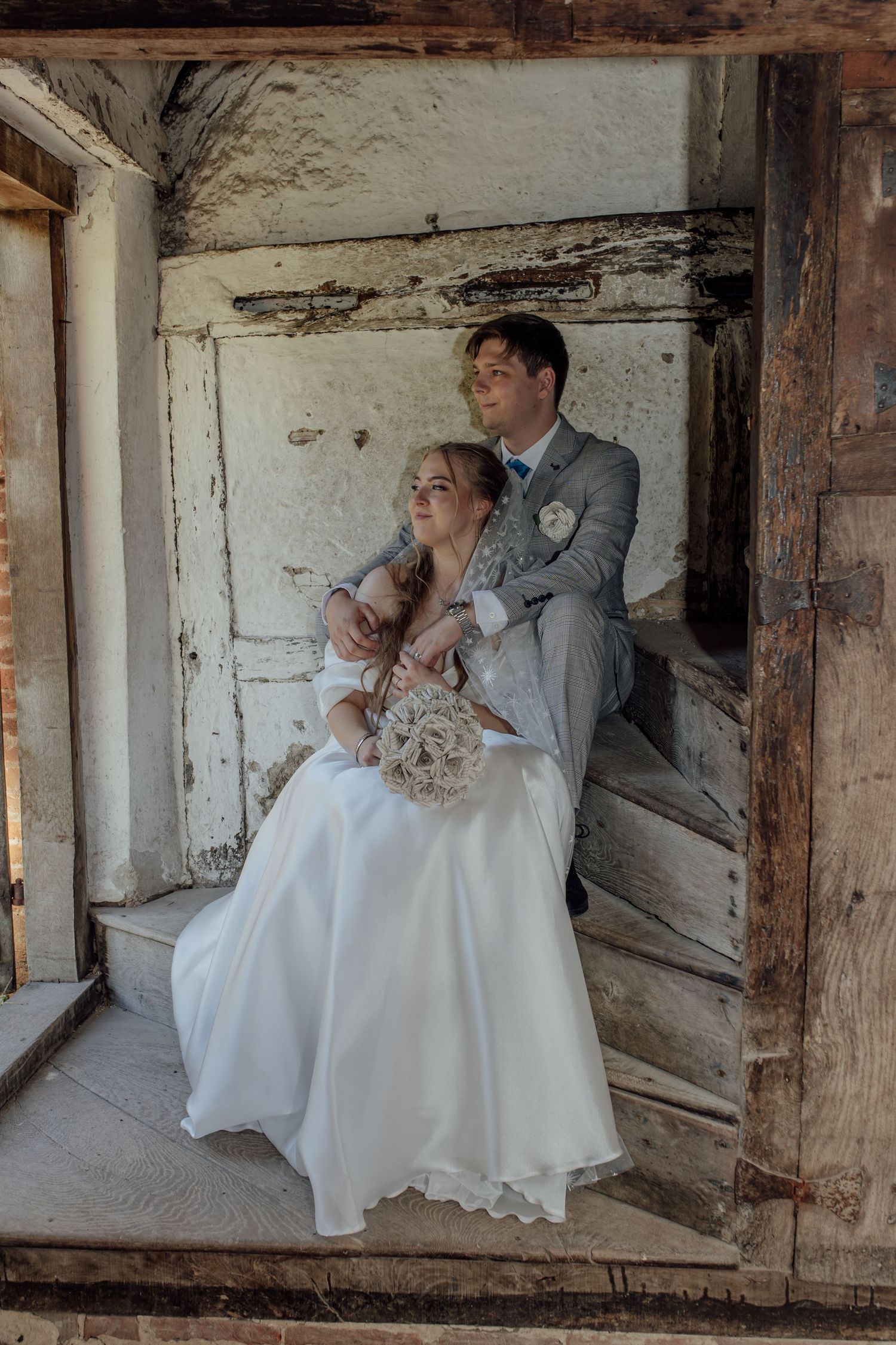 A couple in wedding attire share an intimate moment in a rustic barn setting with exposed wooden beams and white walls.