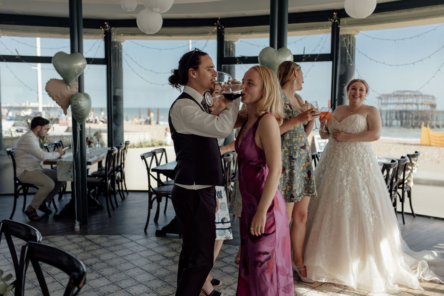 Wedding guests dance together during a reception celebration in an elegant venue with large windows and white paper lanterns.