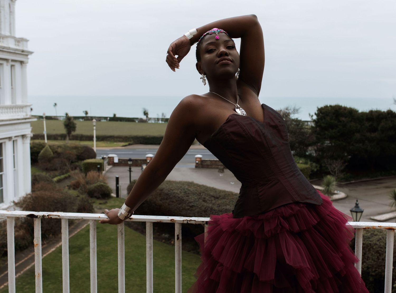 A person in a burgundy corset and tulle skirt poses dramatically on a balcony overlooking coastal scenery.