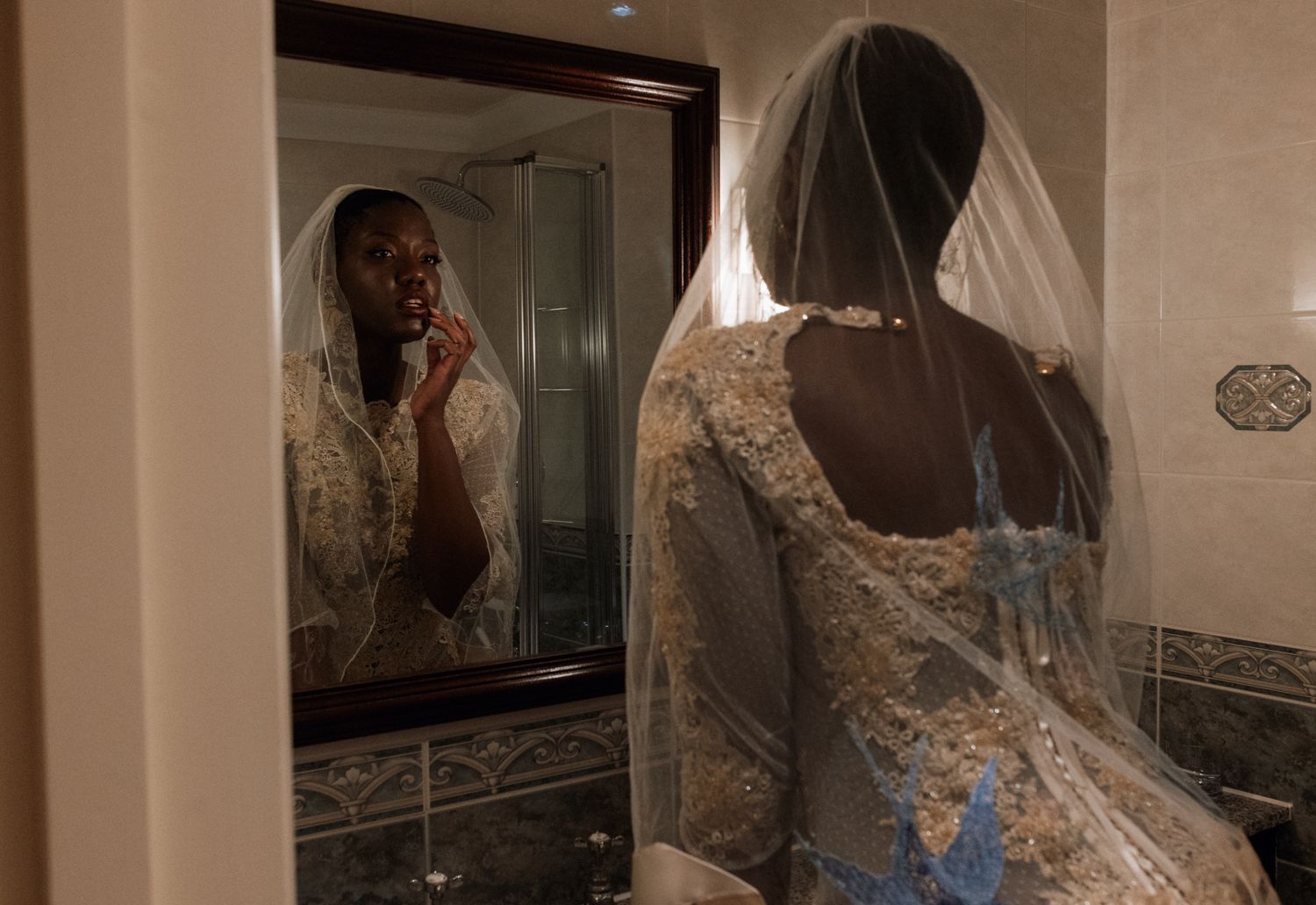 Someone in an elegant lace wedding dress looks into a bathroom mirror with a delicate veil hanging down their back.