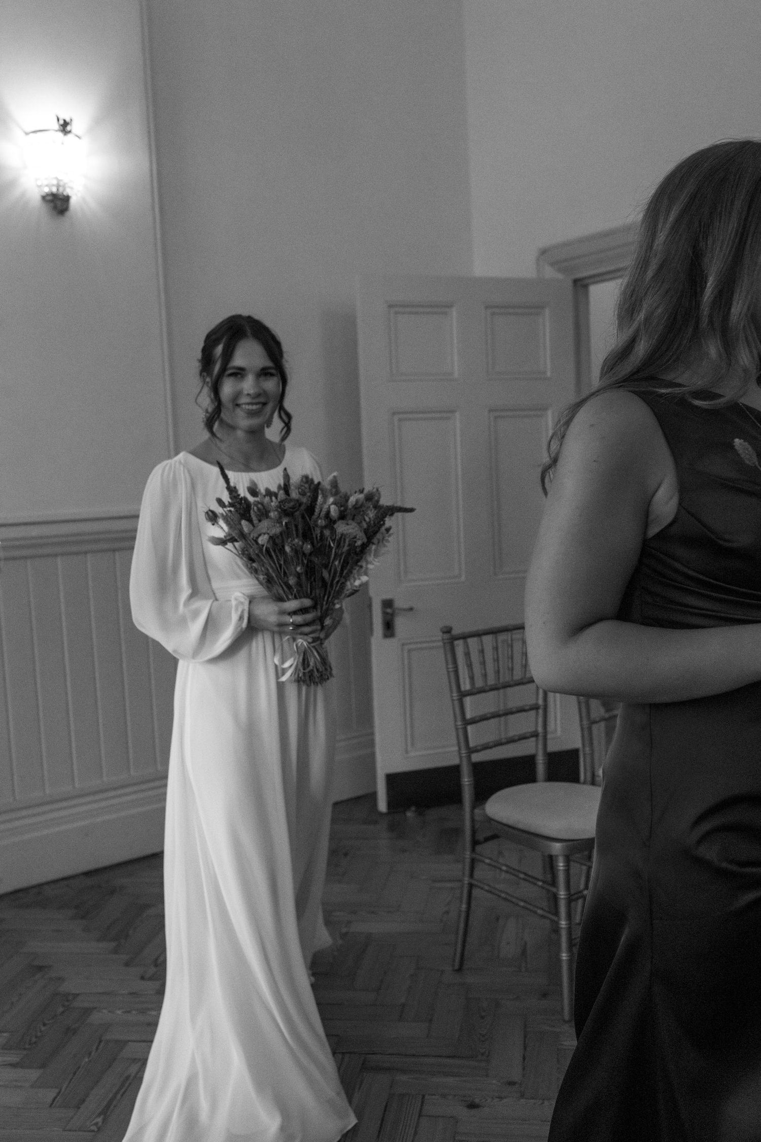 A bride in a flowing white gown holds a bouquet while standing in an elegant room with parquet flooring and white walls.