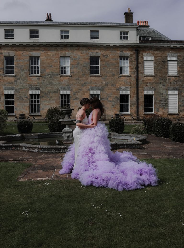Two people embrace in front of a historic stone mansion wearing a stunning purple tulle wedding gown.