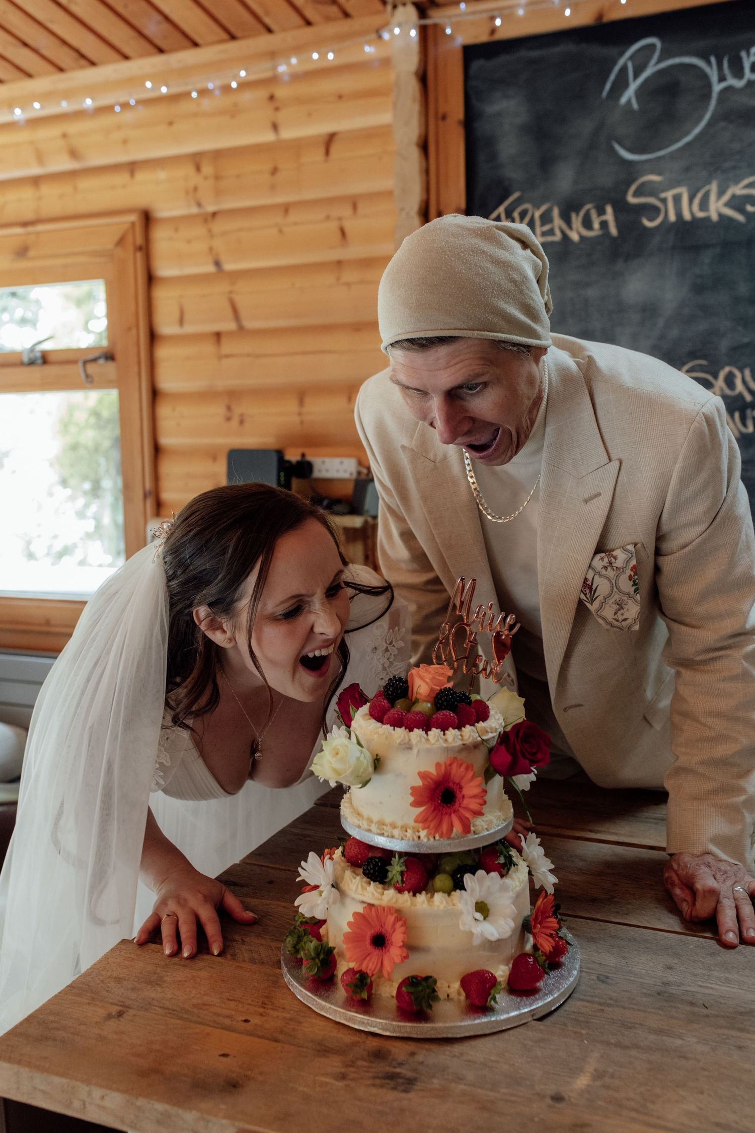 A couple shares a playful moment biting their wedding cake together in a rustic wooden venue decorated with flowers.