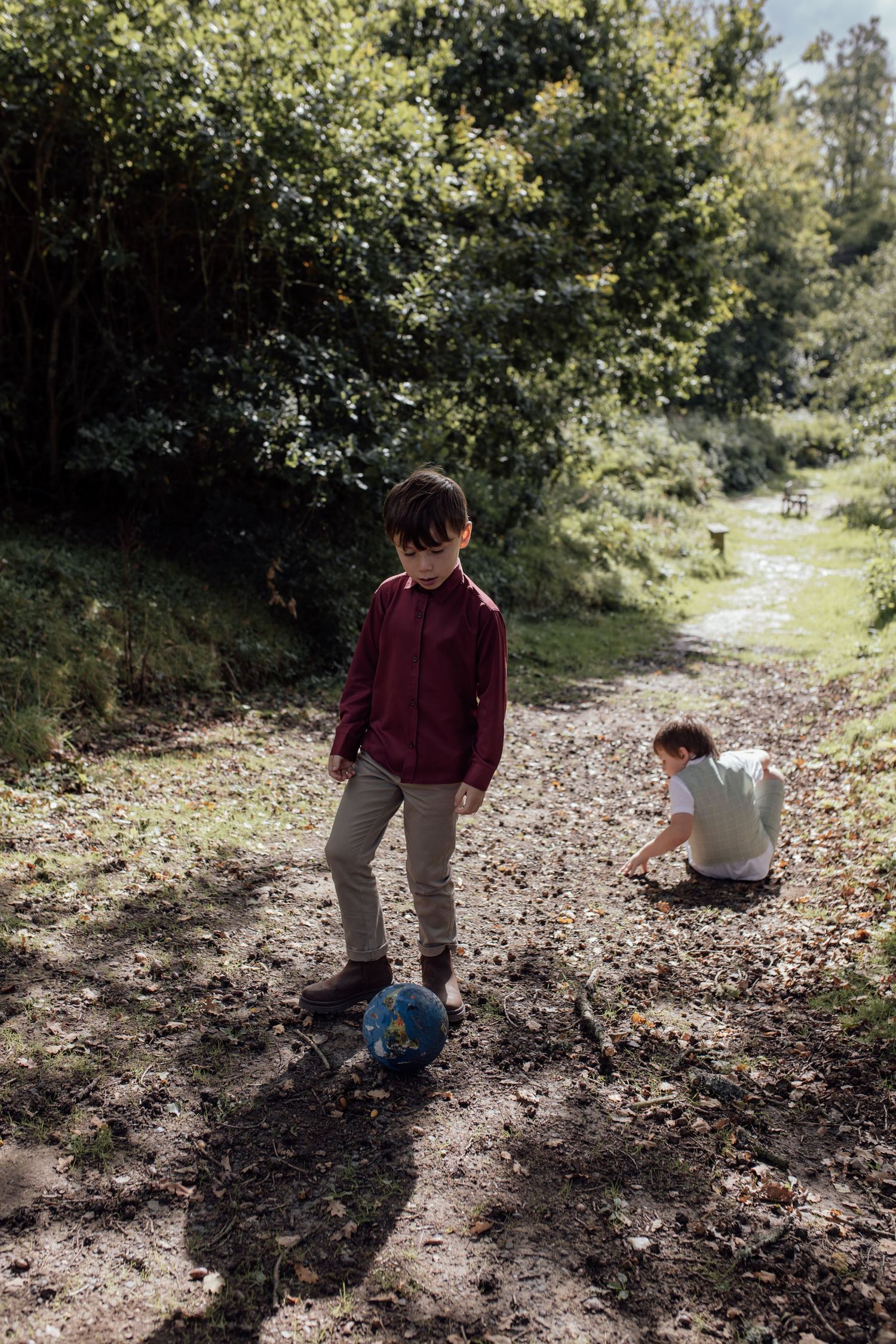 Two people sit on a leaf-covered forest path on a sunny autumn day, casting long shadows through the trees.