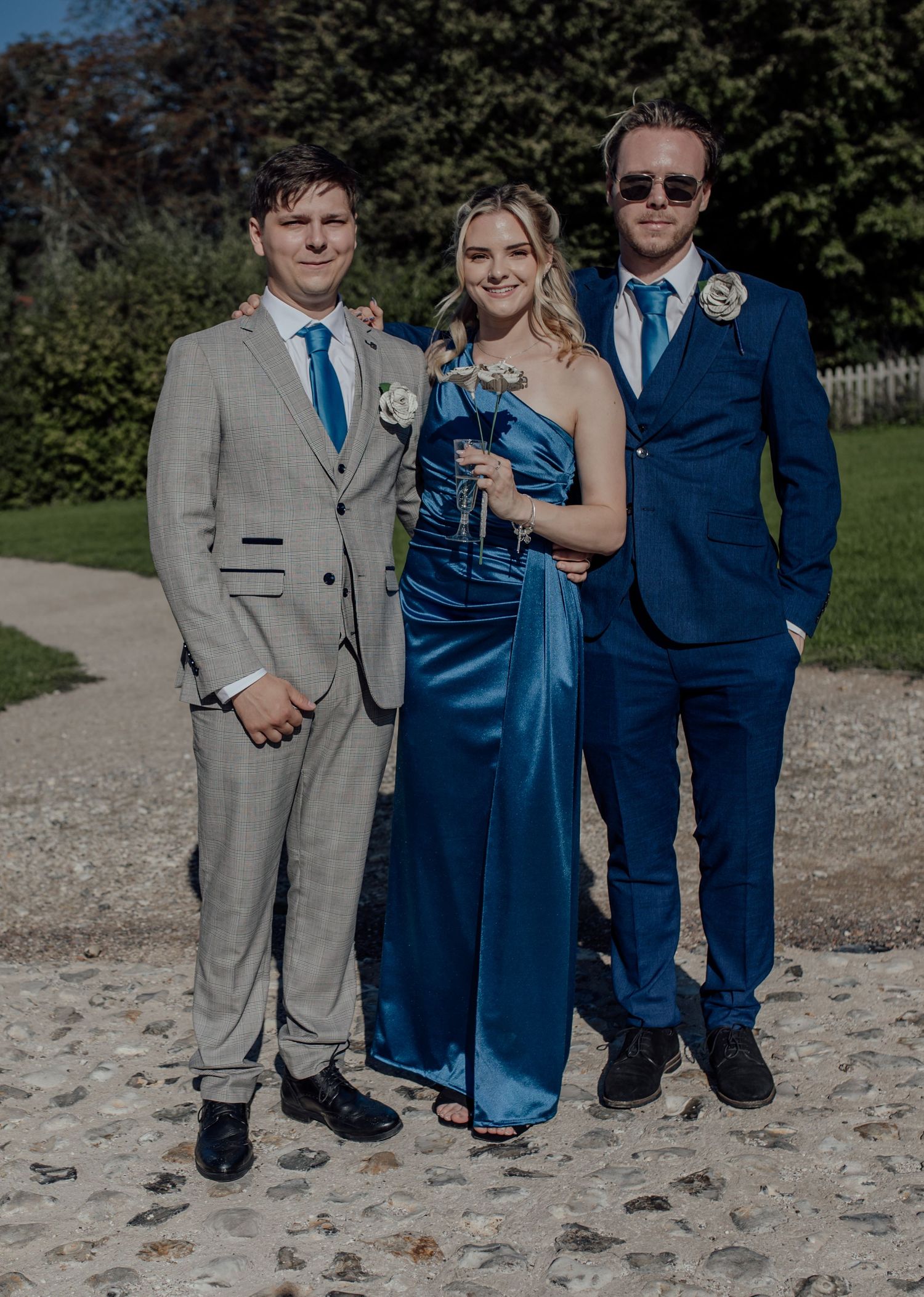 Three people in formal attire stand together outdoors on a sandy surface wearing navy blue and gray suits.