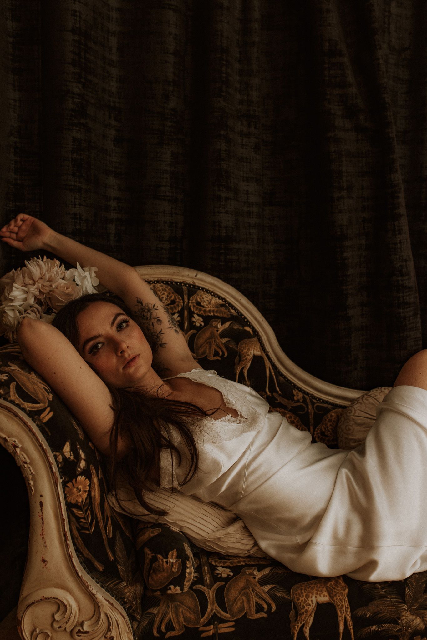 Dramatic boudoir photo of person in white dress reclining on ornate vintage chaise lounge.
