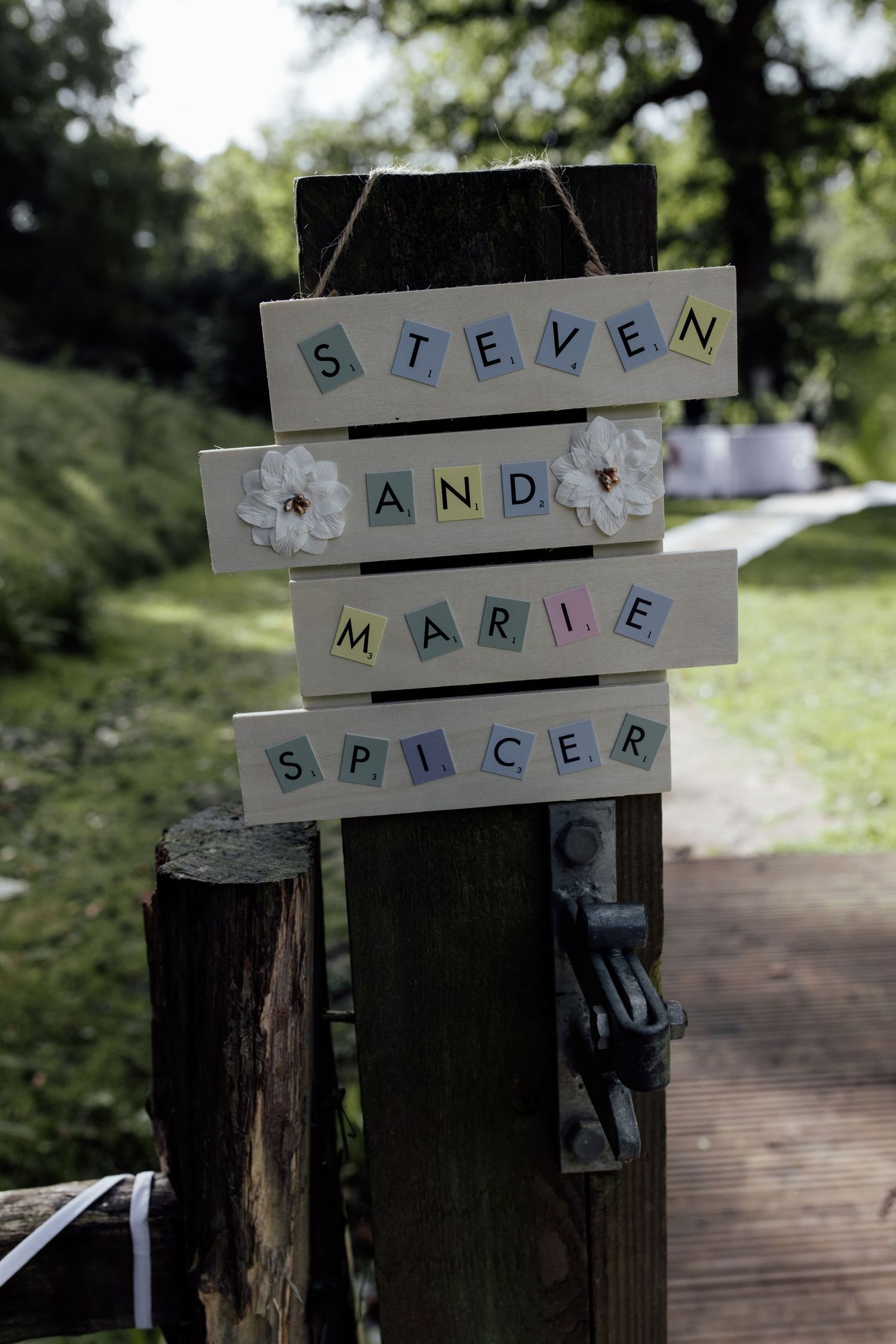A rustic wooden sign displays names 'Steven and Marie Spicer' spelled out in blue and gray letter tiles against a garden background.