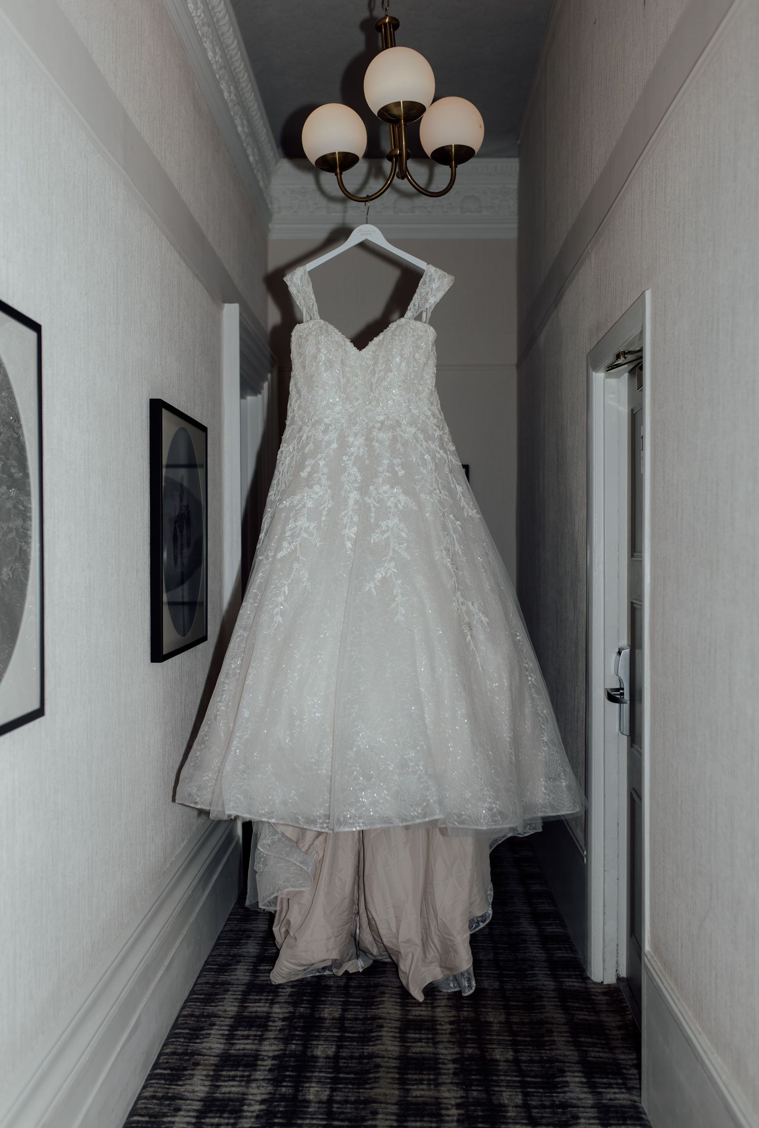 A white wedding dress hangs in a dimly lit hallway with patterned carpet and chandelier lighting.